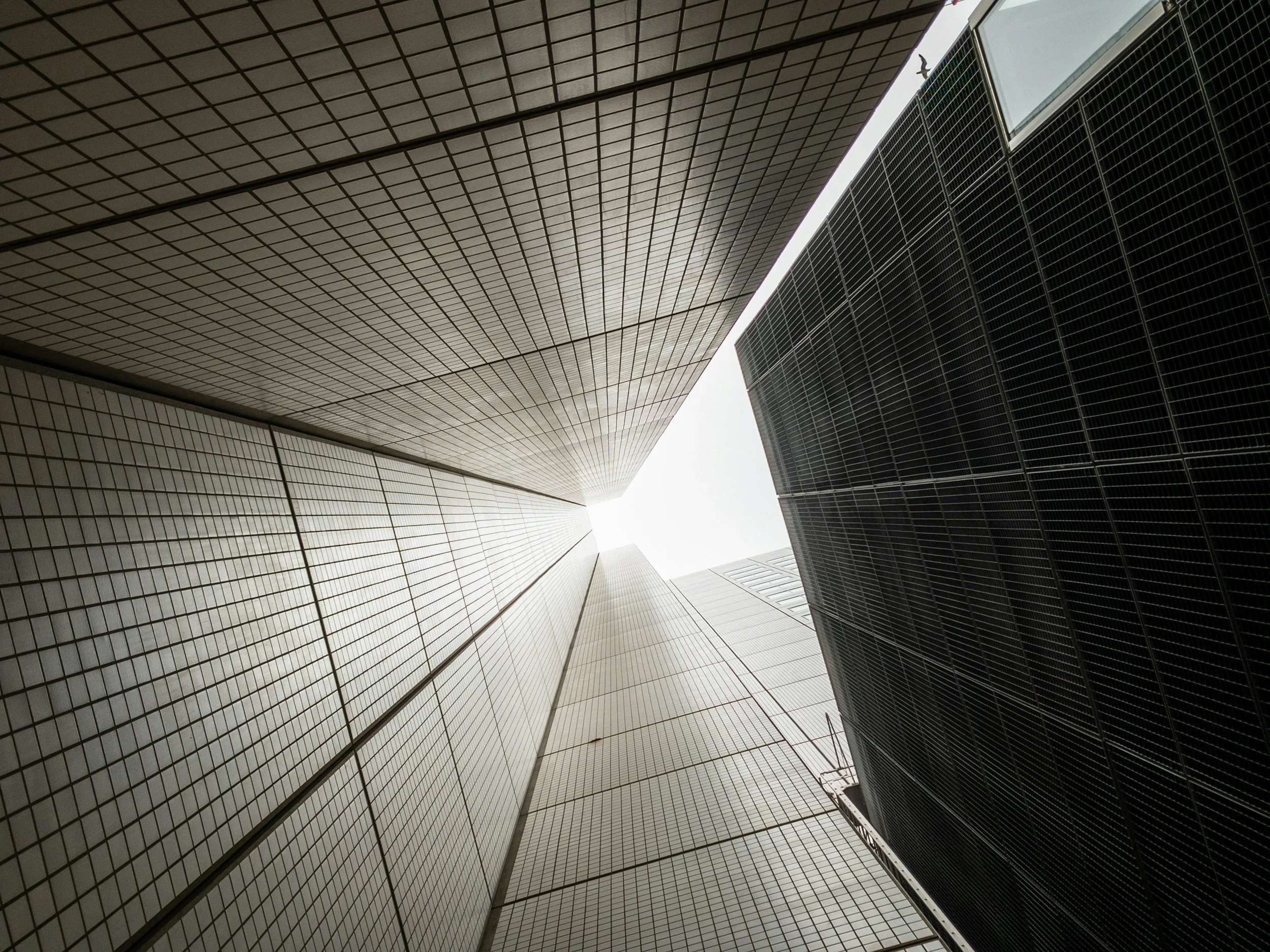 Looking up between tall, modern skyscrapers against a bright sky, with one building covered in white tiles and the other with dark glass panels.