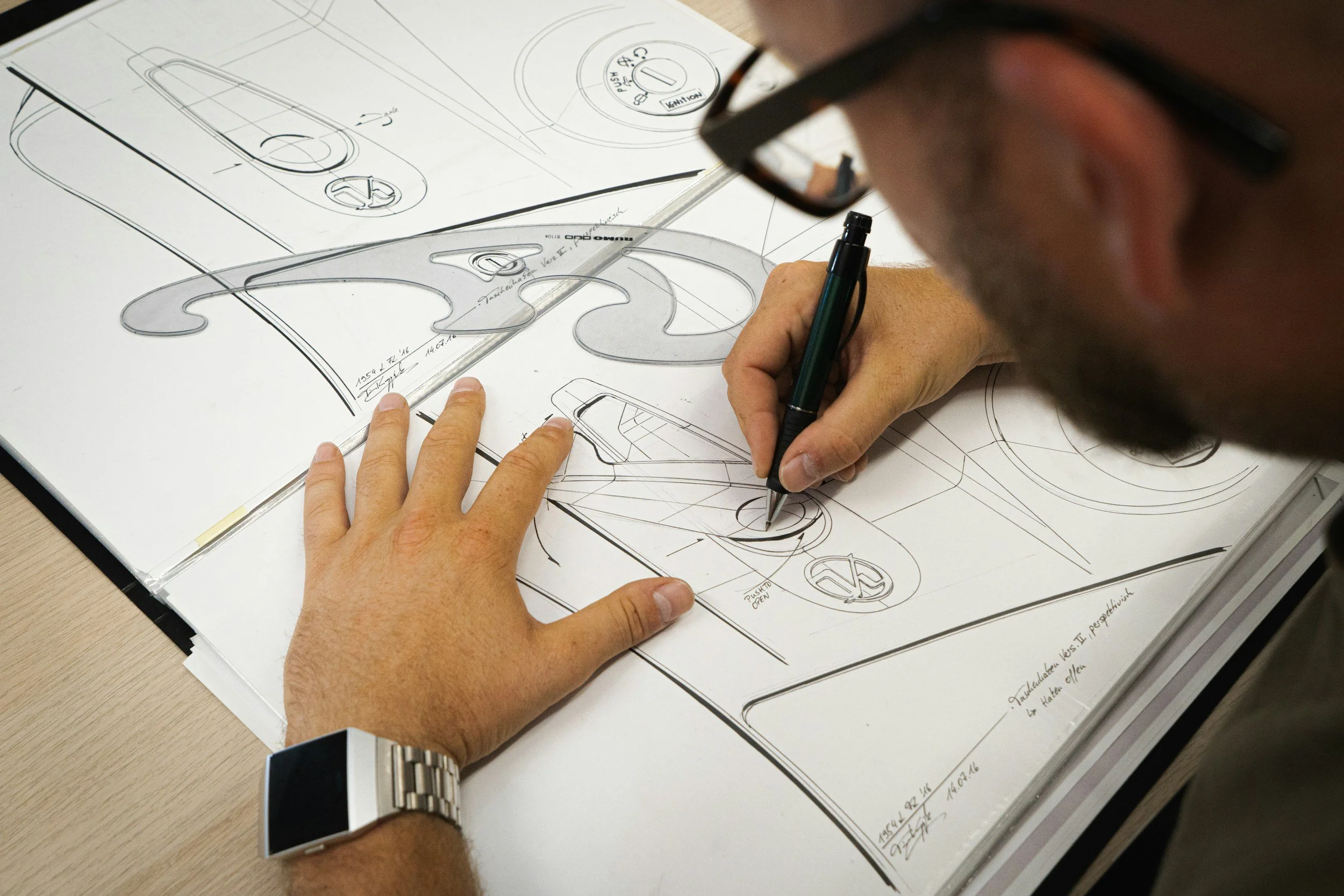 A person with glasses and a watch is working on a technical blueprint of a car design, using a pen and a ruler on a table.