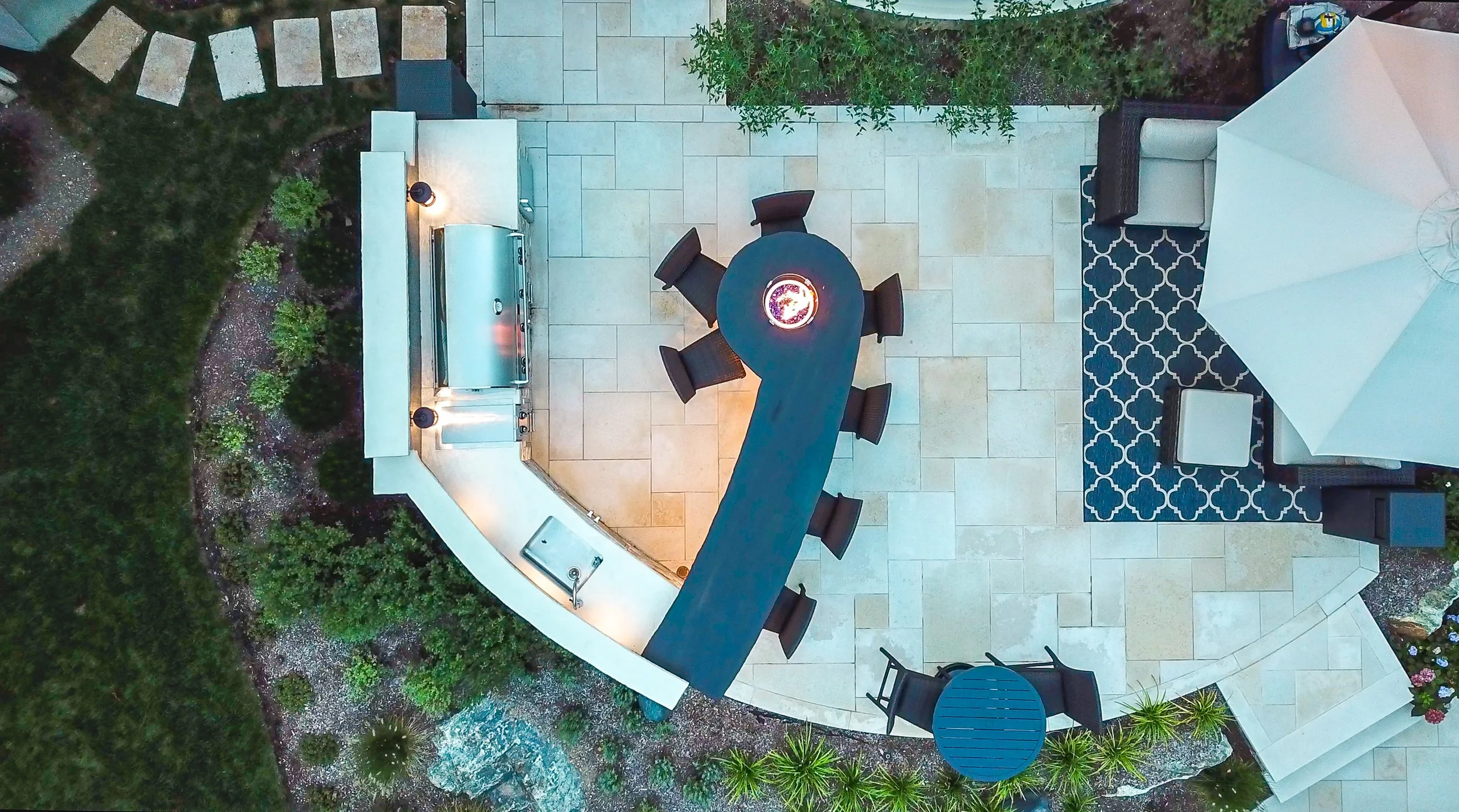 Aerial view of an outdoor patio featuring a curved white outdoor kitchen with a built-in grill, a bar-height black table with six chairs, a white outdoor sofa with an umbrella, and a small black side table, surrounded by plants and decorative paving stones.