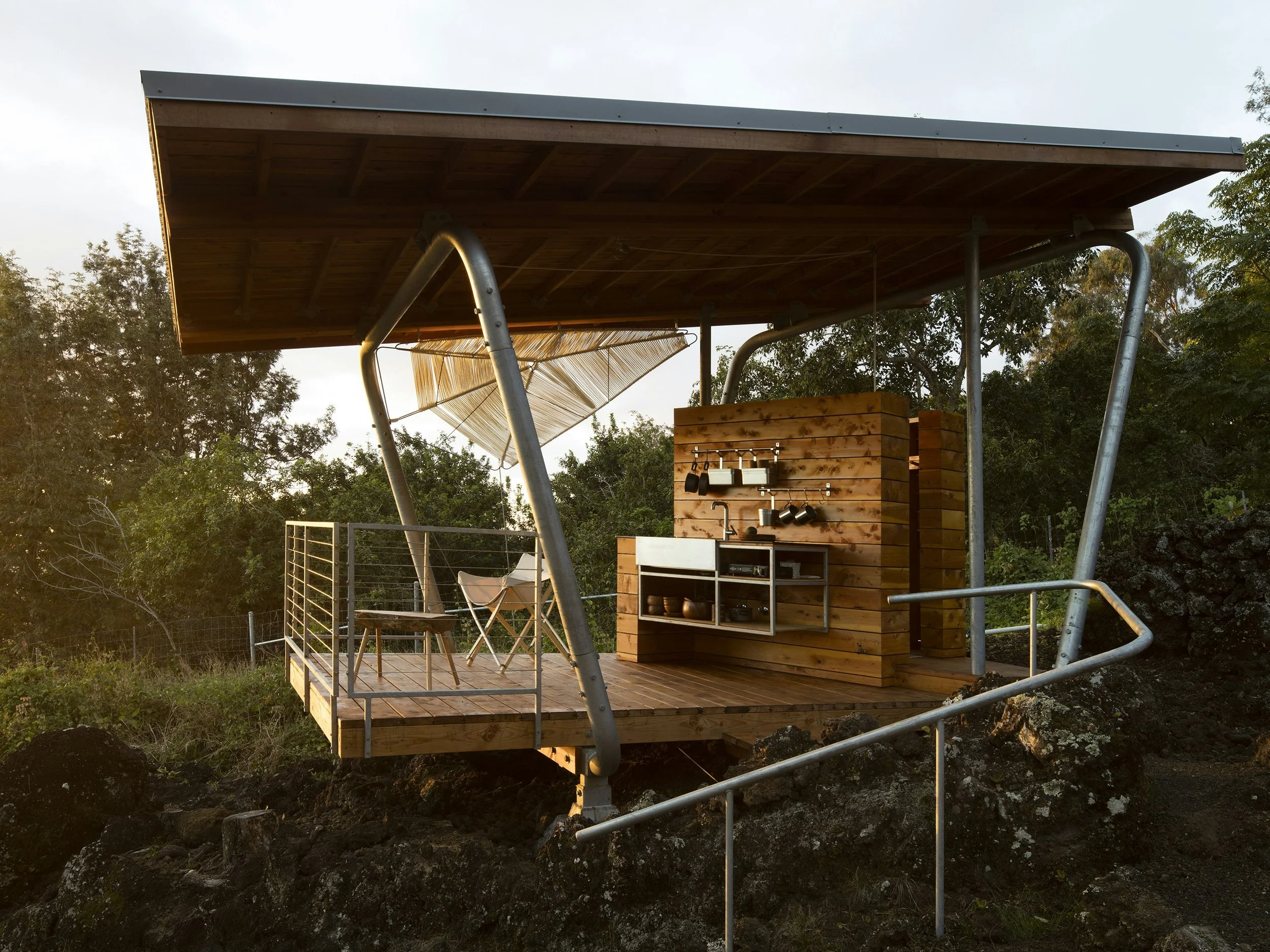 A small outdoor wooden deck with a kitchen setup, chairs, and railing, located on rocky terrain surrounded by trees.