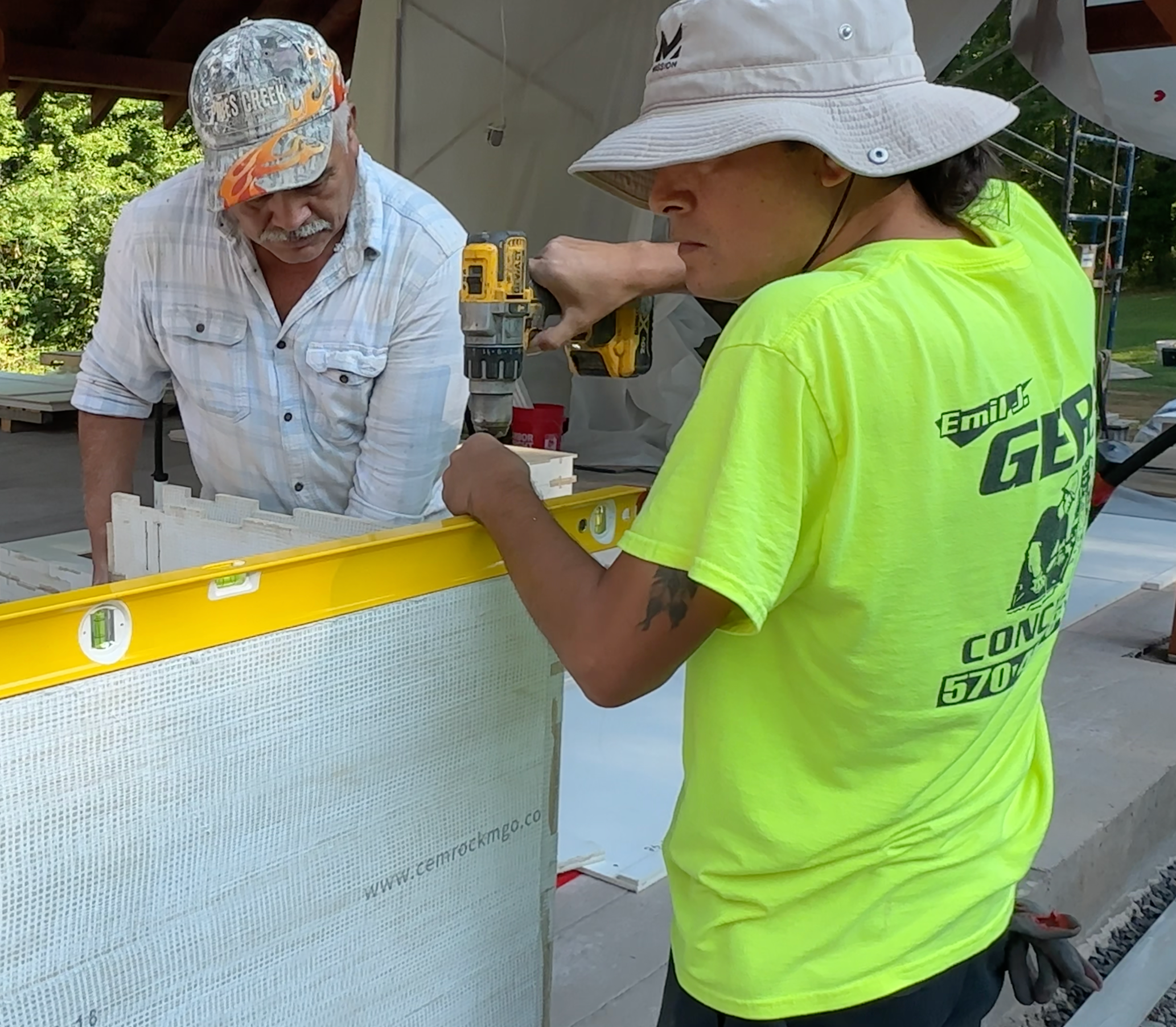 Two men working on construction, one wearing a white hat and the other wearing a gray hat, using a drill on a yellow and white panel.
