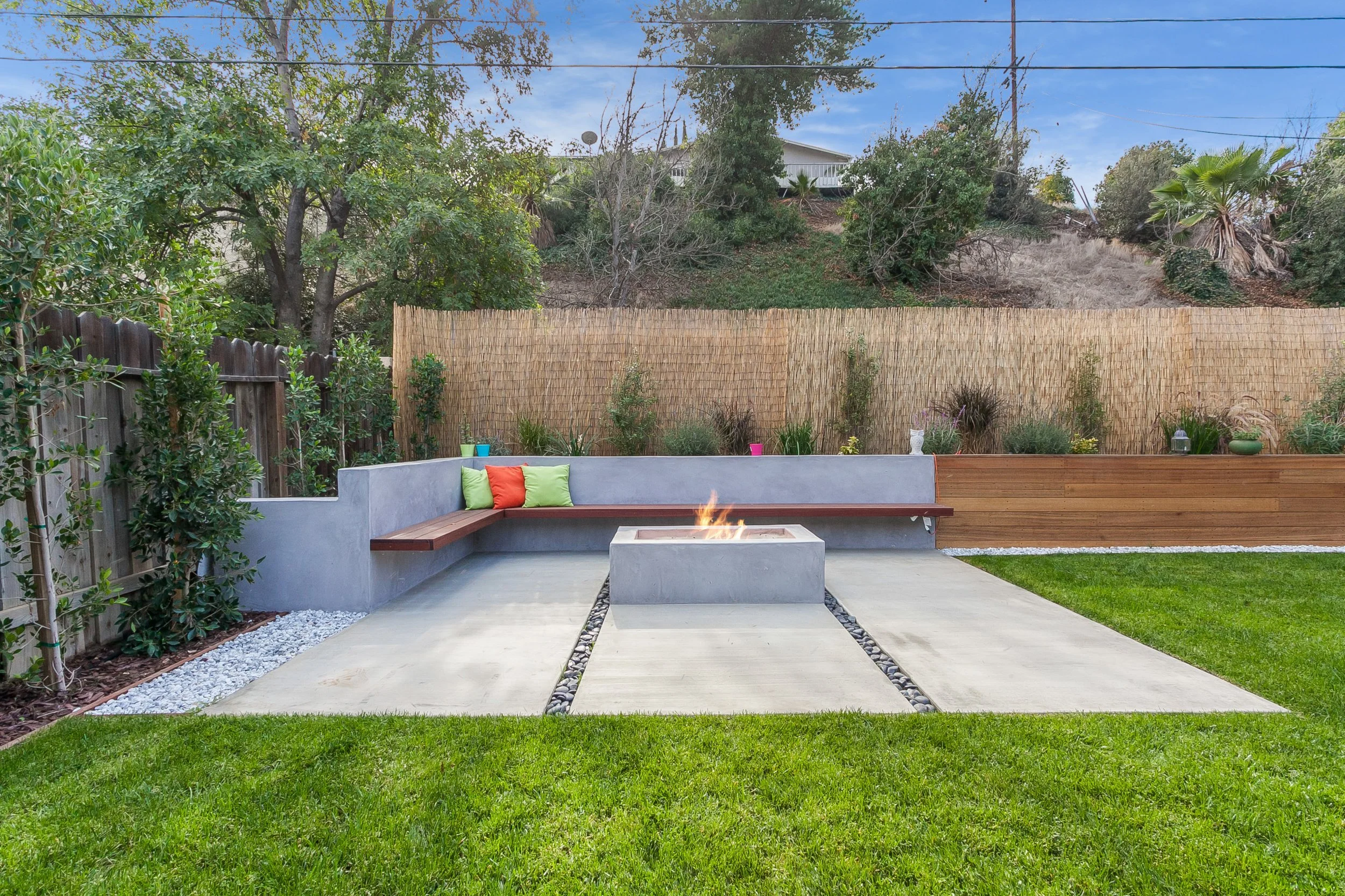 Backyard with concrete patio, built-in concrete fire pit with flames, surrounding grassy yard, wooden and bamboo fencing, trees, and potted plants.