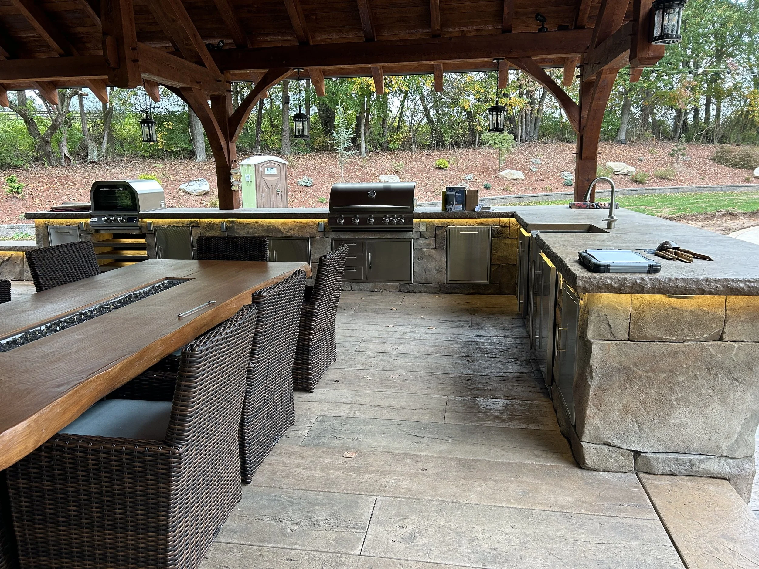 Outdoor kitchen with a stone counter, grill, and seating area under a wooden pavilion, overlooking a landscaped yard with trees and rocks.