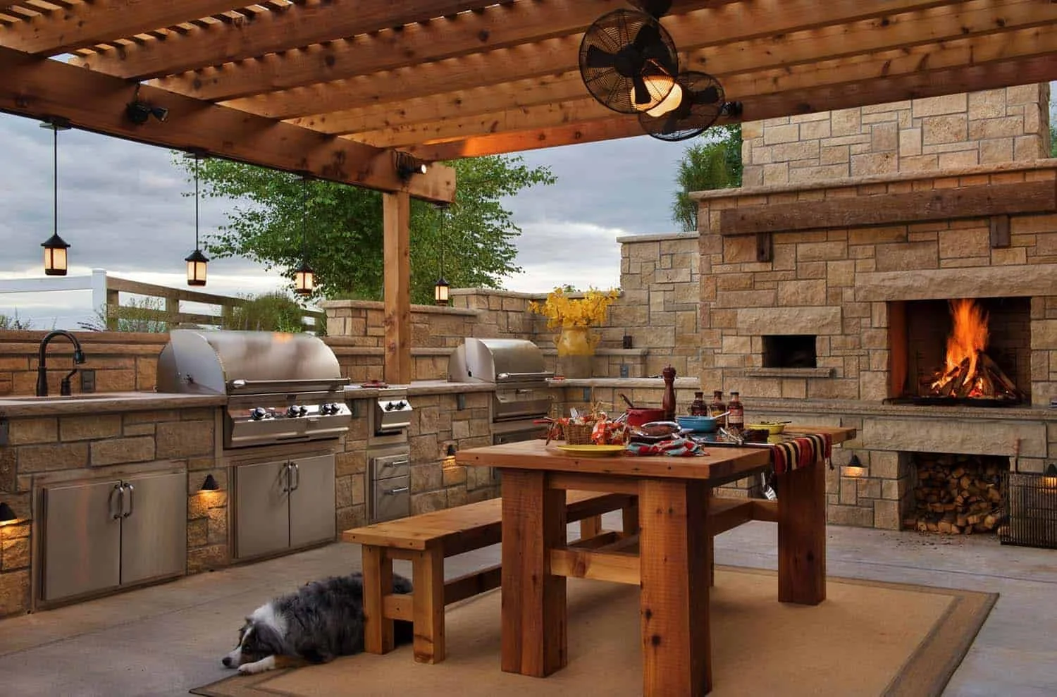 Outdoor kitchen with stone fireplace and wood ceiling, stainless steel grills, wooden dining table, and a dog lying on the floor.