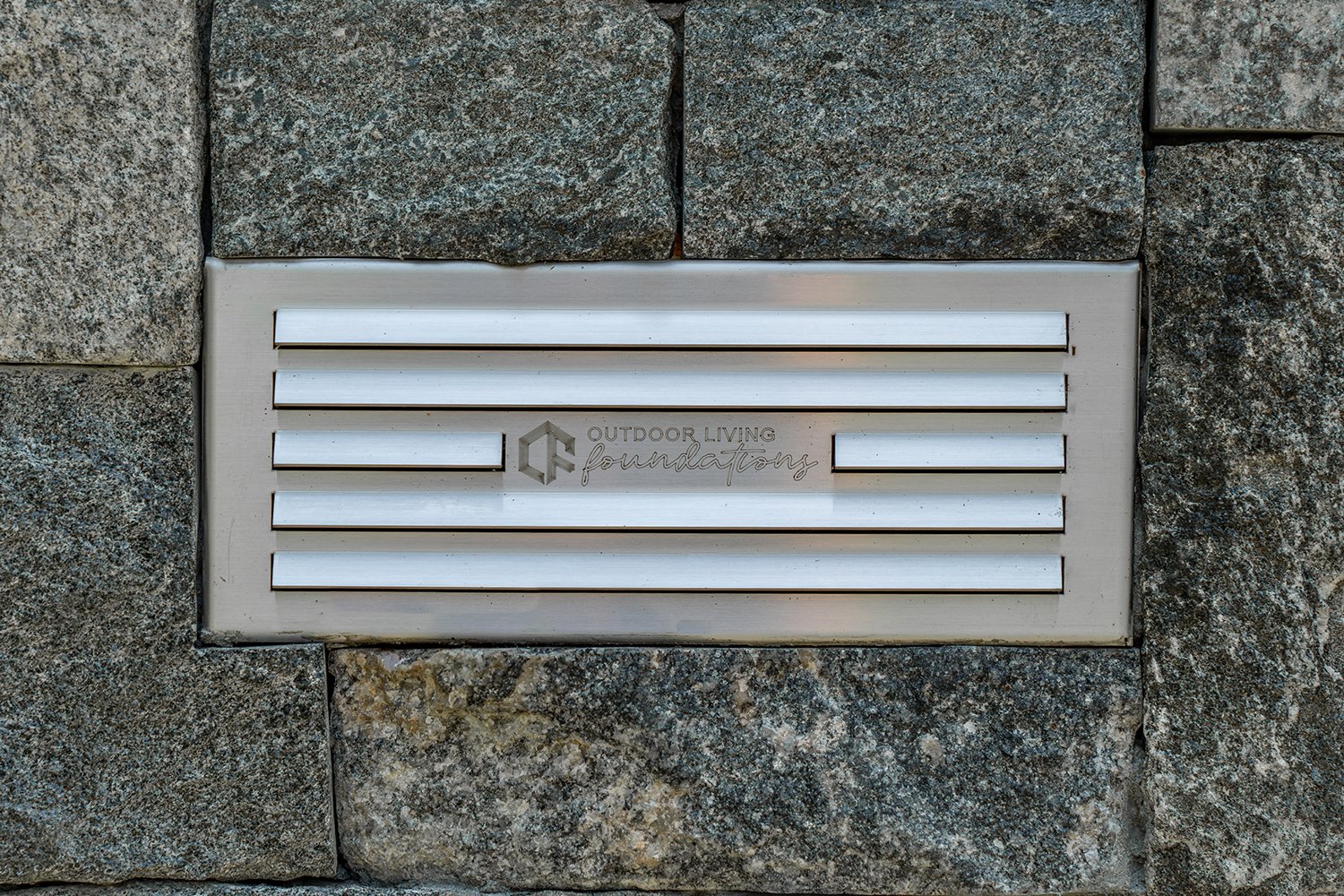 Close-up of stainless steel outdoor kitchen vent panel with engraved Outdoor Living Foundations branding, framed by durable stonework. Essential for proper ventilation and safe outdoor kitchen construction. outdoor kitchen ventilation, stainless stee
