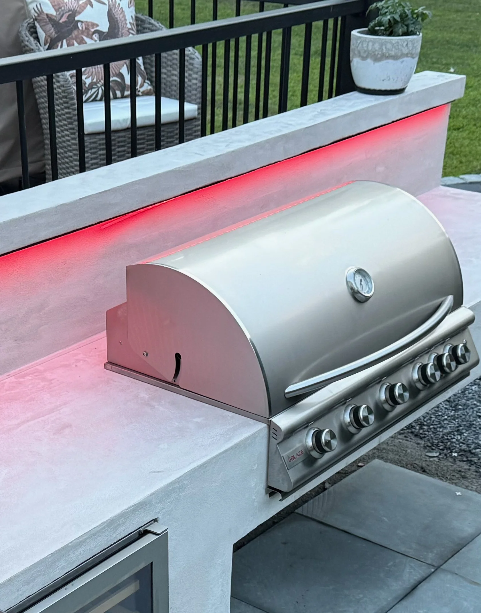 Outdoor kitchen grill with stainless steel lid and control knobs, red LED lighting along the backsplash, and a decorative plant on a concrete countertop.