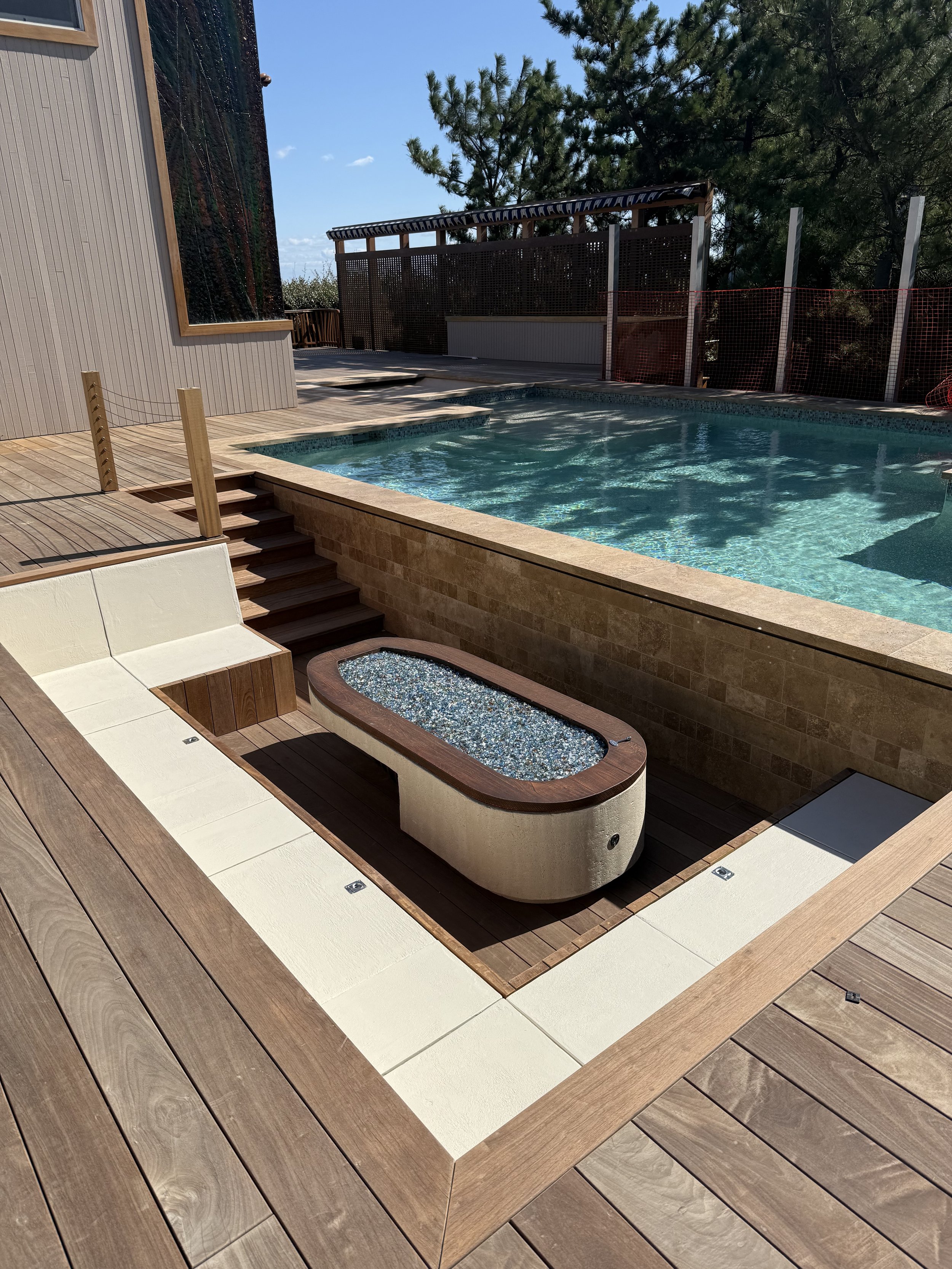 A backyard with a swimming pool, a hot tub filled with water, a wooden deck, and a fence with trees in the background.