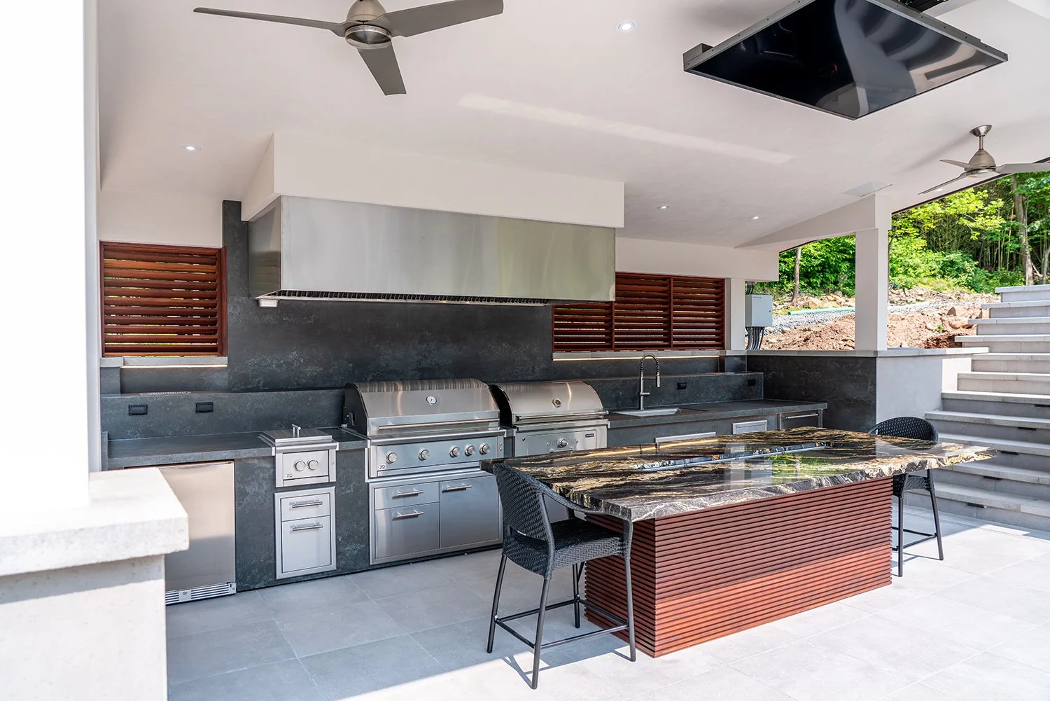 Luxury covered outdoor kitchen featuring dual stainless steel grills, a sink, under-counter appliances, and a polished stone island bar. Complete with ceiling fans, modern lighting, and a mounted TV for the ultimate backyard entertainment hub.
covere