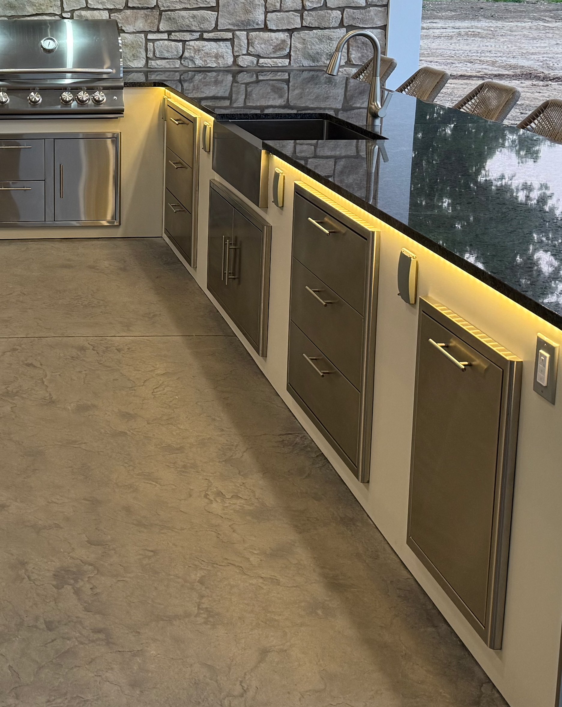Modern kitchen with stainless steel appliances, stone wall, black countertop with inlaid lighting, and bar seating with woven chairs.
