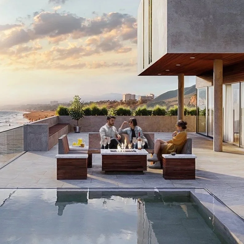 Three people sitting on outdoor patio furniture near a fire pit with drinks, overlook a beach at sunset.