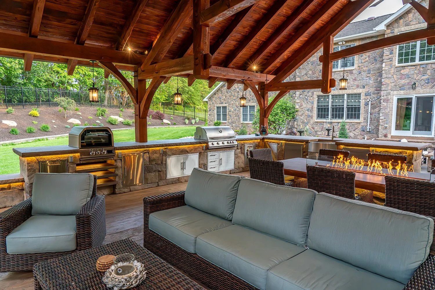 Built By Emil Gera. Stunning rustic outdoor kitchen with a wood-beam pavilion, stone countertops, stainless steel built-in grills, and a dining fire table. This backyard entertaining space blends luxury and comfort with cozy wicker seating and warm l