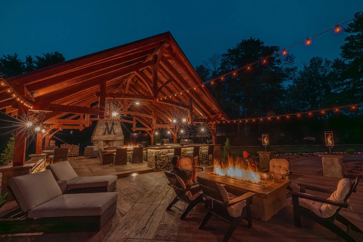 Built By Emil Gera. Cozy outdoor pavilion kitchen with timber frame beams, stone fireplace, fire table, and string lighting for evening entertaining. Includes a full outdoor kitchen with grill, sink, and stone bar island for a warm and inviting backy