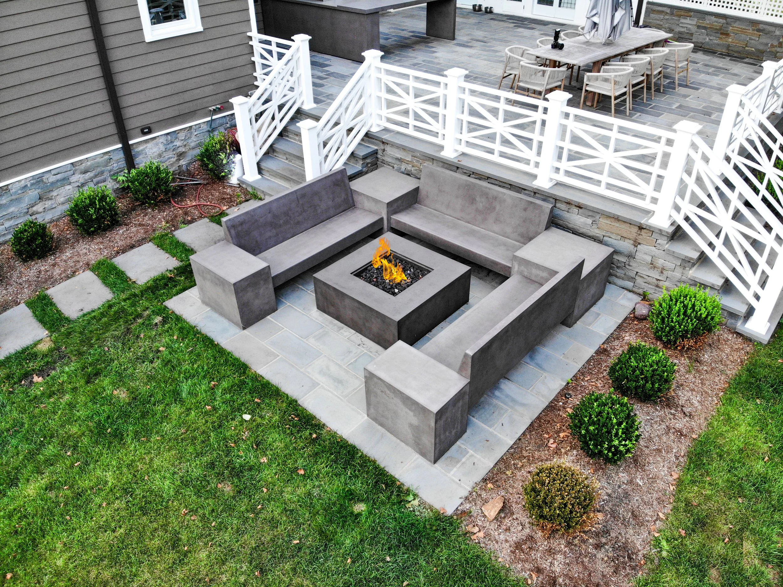 A backyard patio with a fire pit and surrounding modern concrete seats, bordered by a stone wall, with a grassy lawn, small bushes, and a white wooden fence with stairs leading to an upper deck area with outdoor dining furniture.