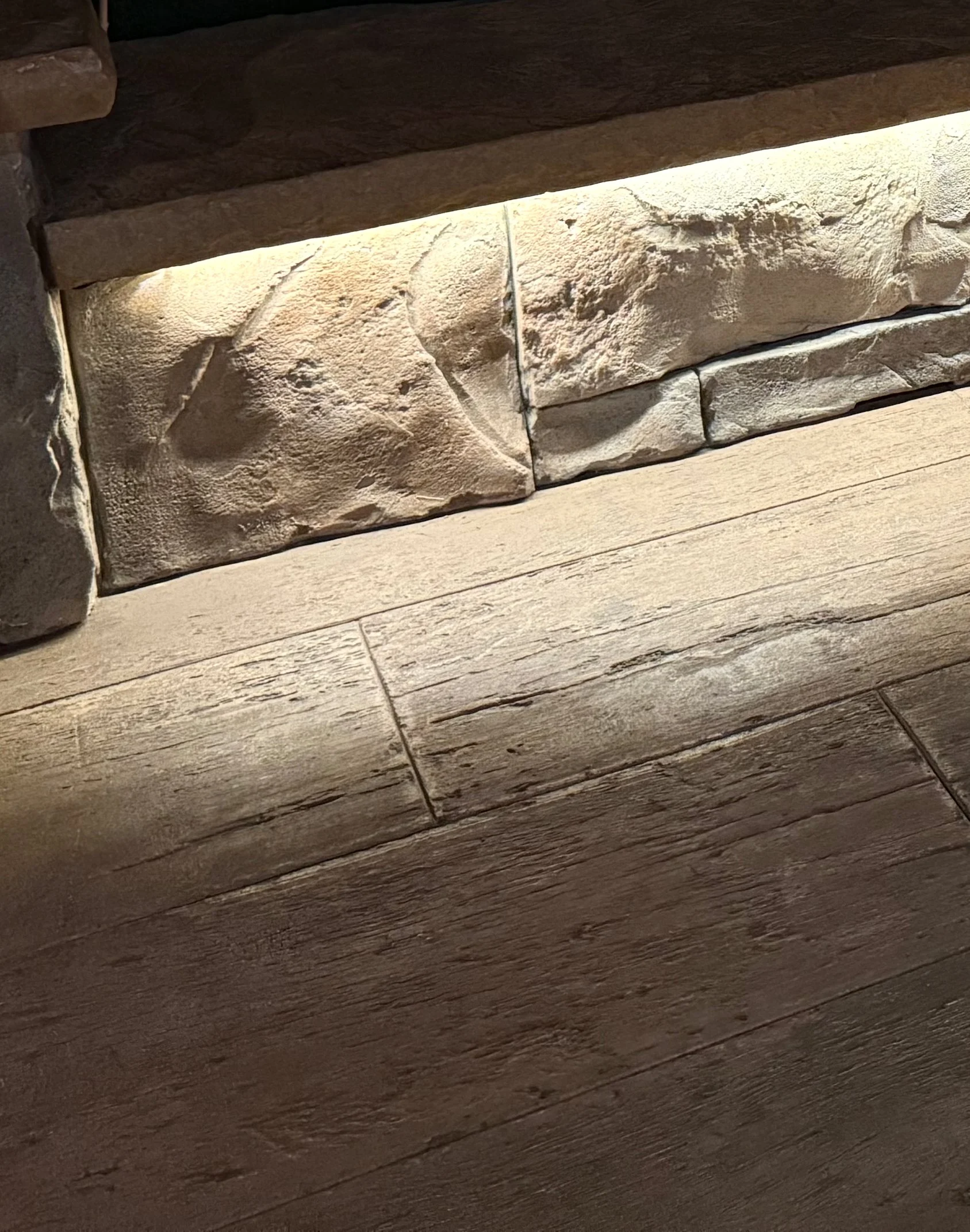 Close-up of a wooden floor with a stone wall in the background, illuminated by a warm light.