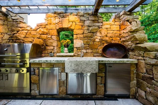 Outdoor kitchen with stone walls, stainless steel appliances, and a concrete countertop with a sink, decorated with a vase of yellow flowers.