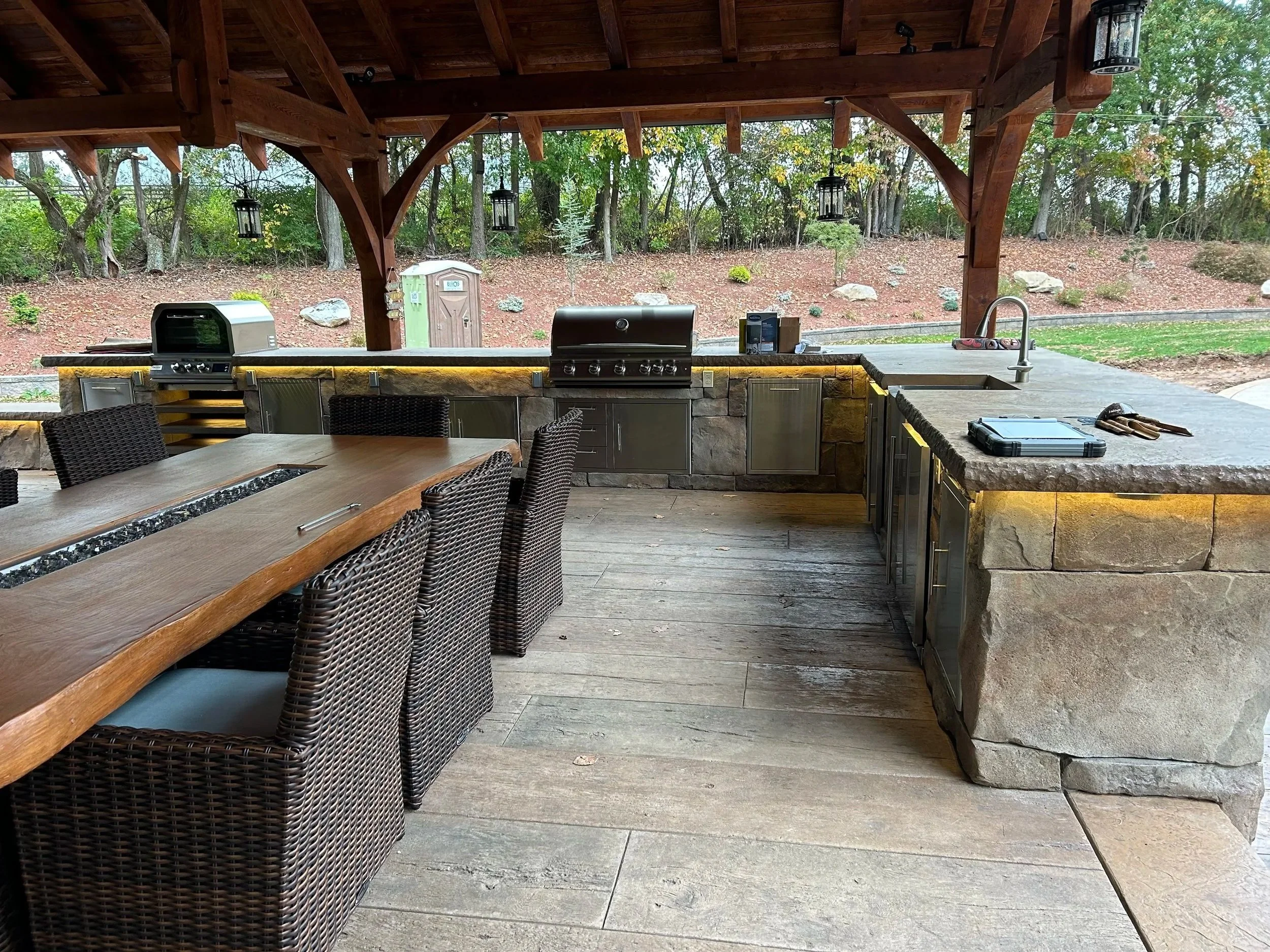 Outdoor kitchen with a stone island, grill, and sink under a wooden pavilion, surrounded by trees and a landscaped yard.