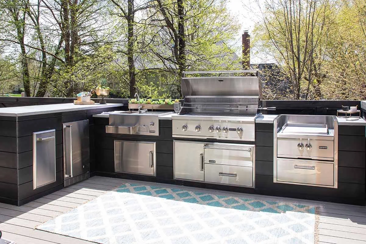 Outdoor kitchen with stainless steel grill, sink, and storage cabinets on a wooden deck, surrounded by trees.