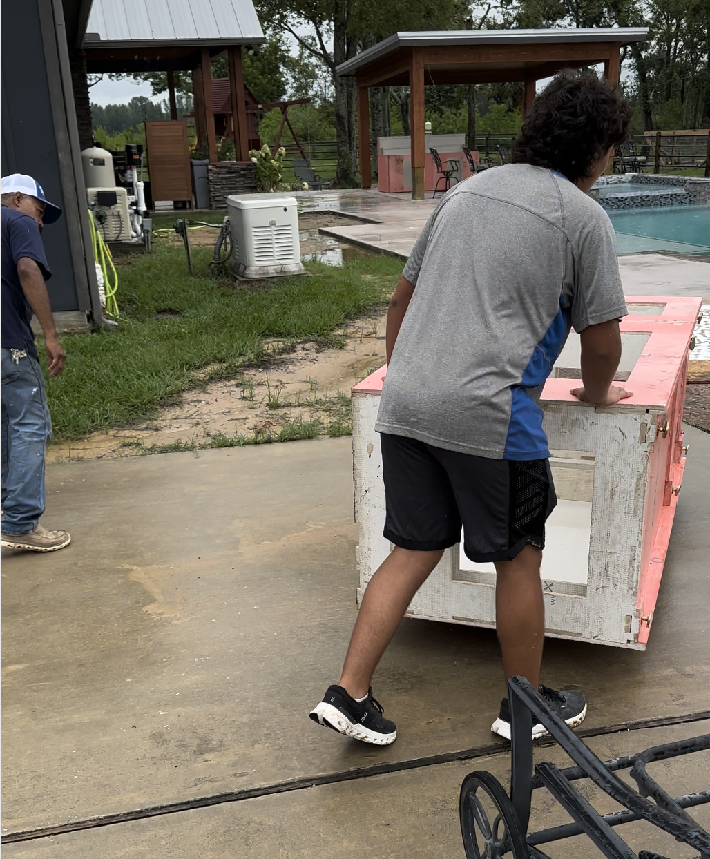 Two people lifting a large, rectangular piece of furniture outdoors near a swimming pool, with a grassy area and wooden structures in the background.