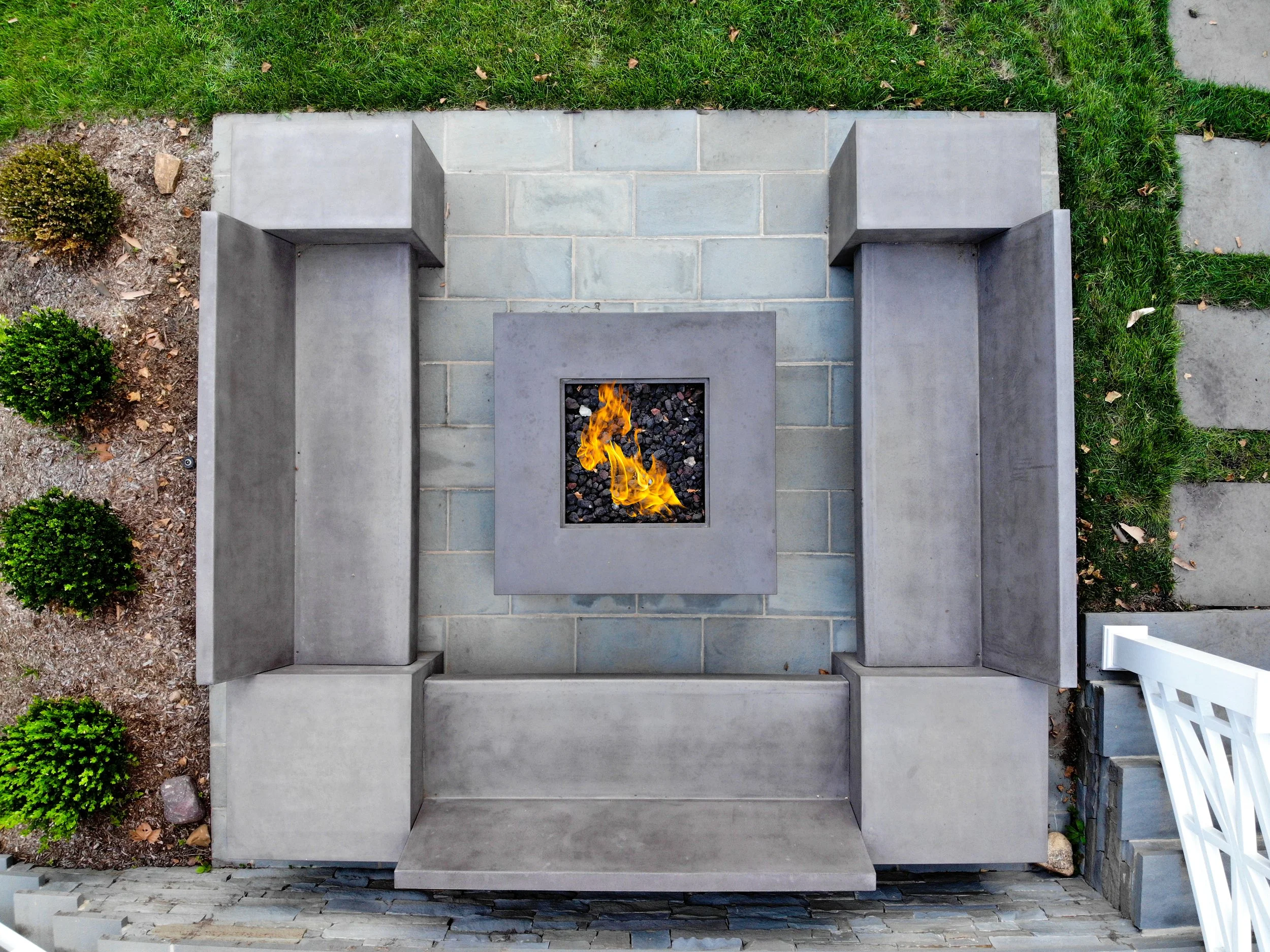 Top-down view of a modern outdoor fire pit with a square flame, surrounded by concrete bench seating on a stone patio, with grass, small shrubs, and stepping stones nearby.