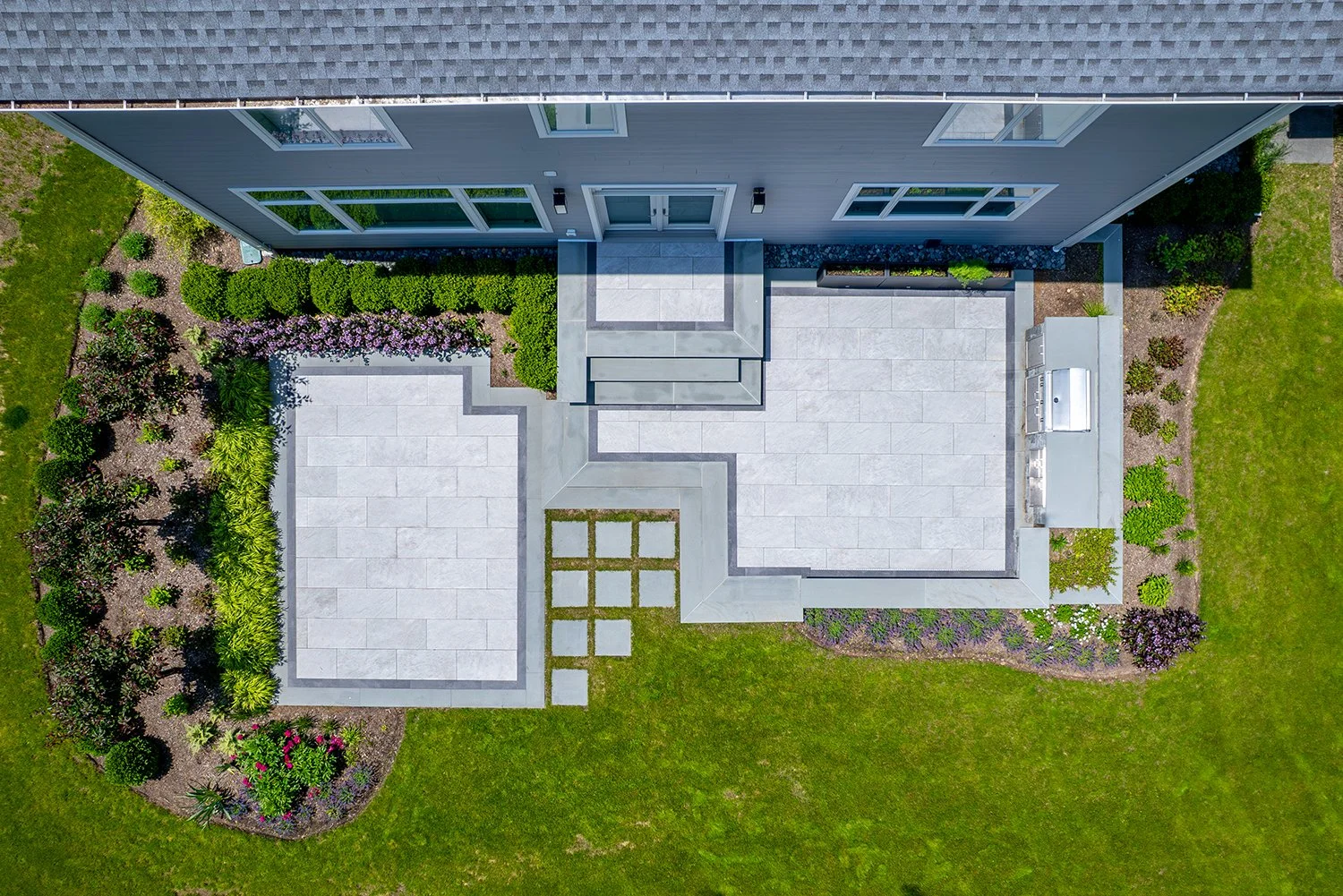 Description: Aerial view of a modern outdoor patio with large stone pavers, geometric hardscape design, and surrounding garden landscaping. The sleek patio layout enhances curb appeal and provides the perfect foundation for an outdoor kitchen or back
