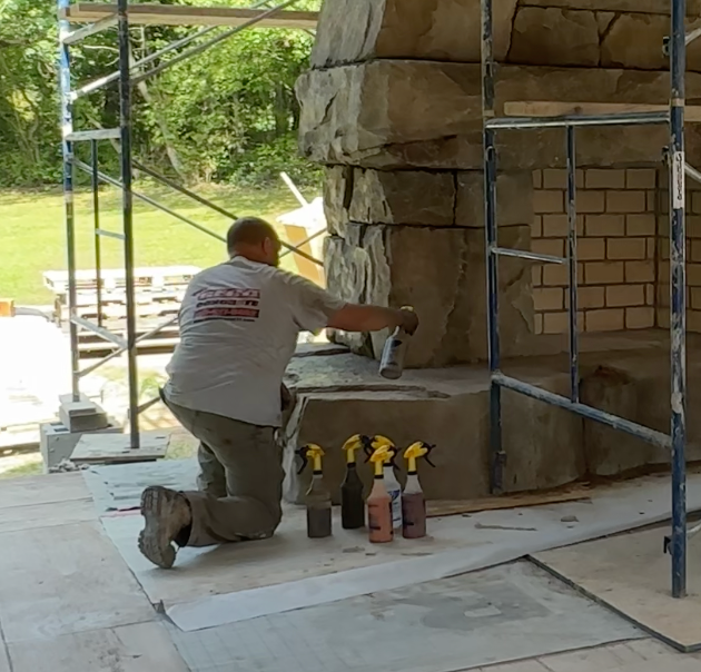 A worker kneeling on the ground applying sealant or adhesive to a large stone in a construction setting with scaffolding around, and three spray bottles placed nearby.