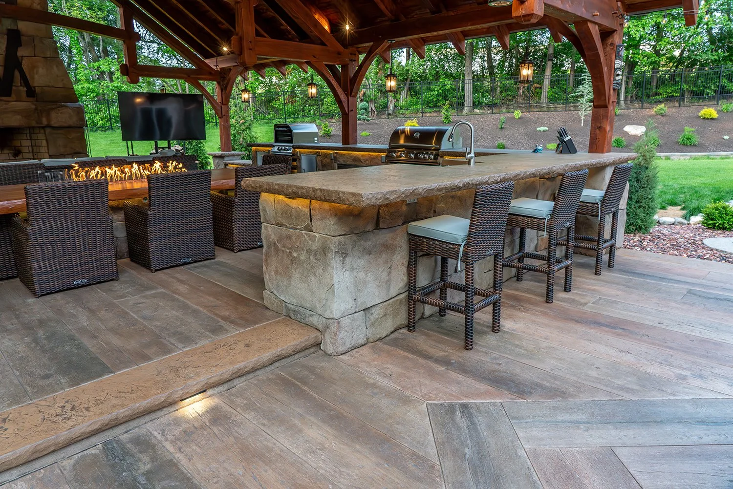 Built By Emil Gera. Rustic-style outdoor kitchen with stone bar island, wicker bar seating, wood beam pergola, and integrated fireplace with mounted TV. Perfect for cozy backyard gatherings and entertaining in all seasons.
 rustic outdoor kitchen, st