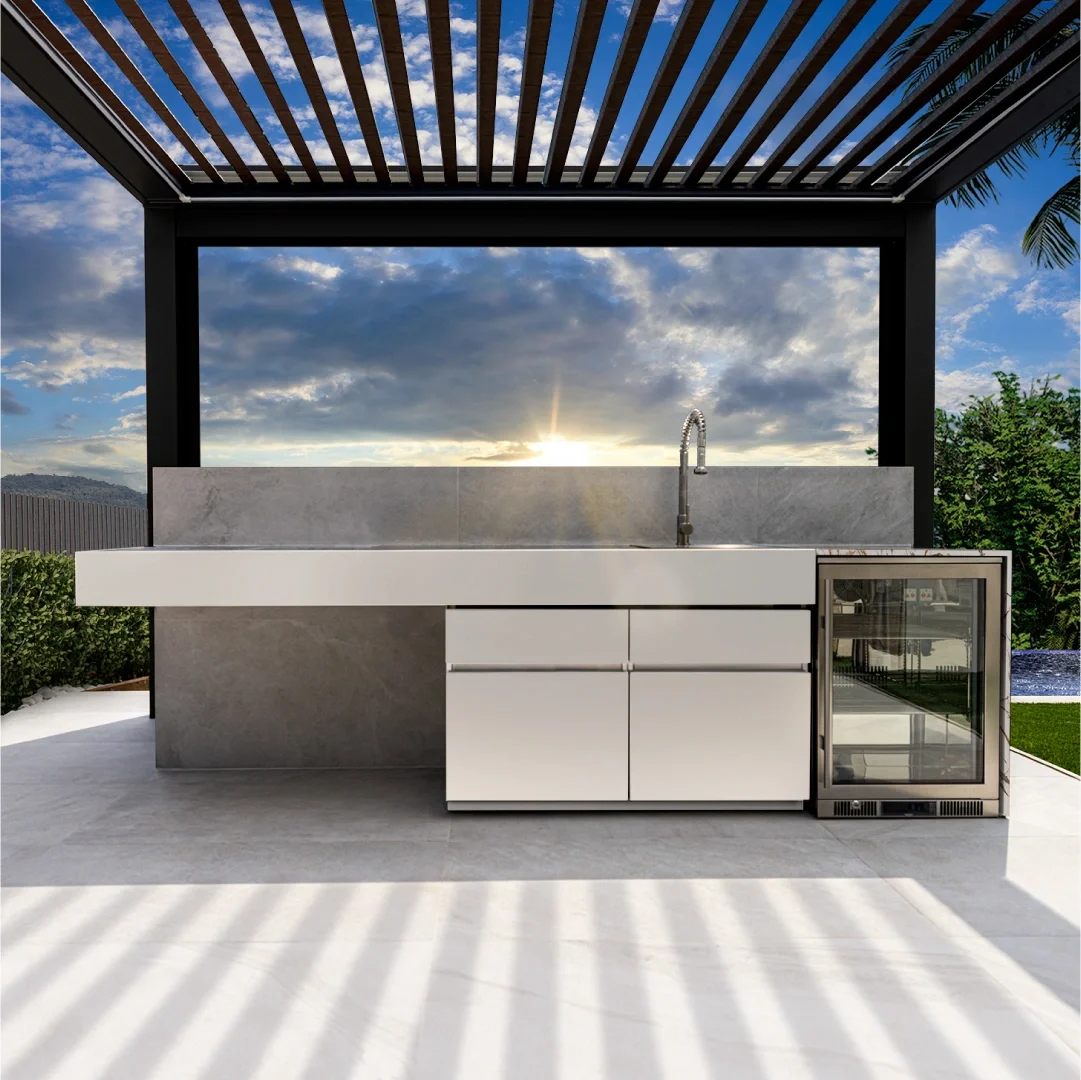 Outdoor kitchen with white cabinetry, a marble countertop and a small refrigerator, under a pergola with slatted roof, overlooking a sunset sky with clouds, trees, and a pool in the background.