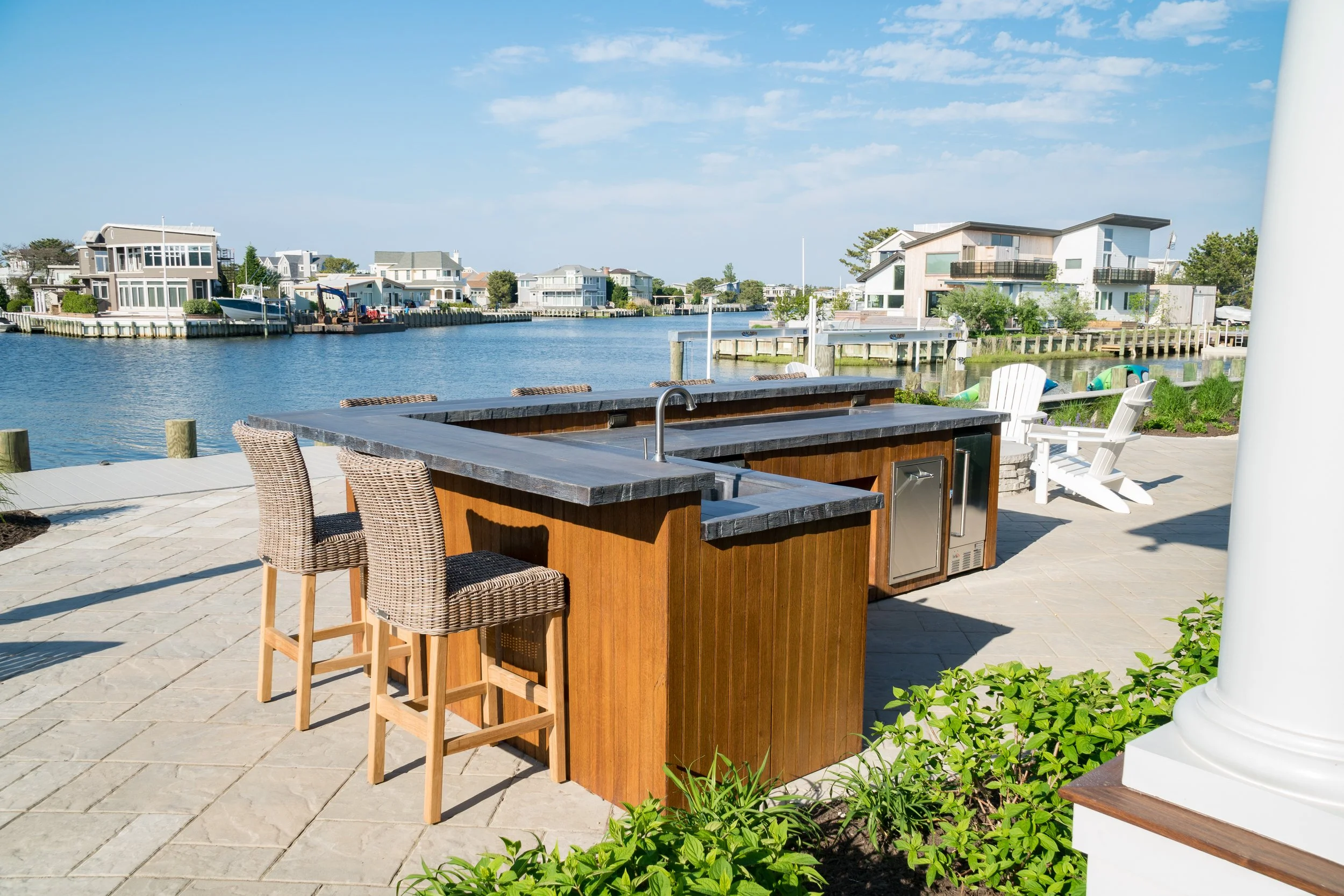 Outdoor bar with a dark wood countertop and wicker chairs, situated near a waterway with modern houses on the opposite side, under a clear blue sky.