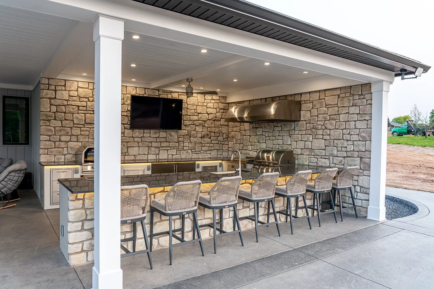 Built By Emil Gera. Elegant covered outdoor kitchen with a long stone bar island, stainless steel appliances, built-in grill, and mounted TV. Stylish bar seating and soft under-counter lighting make this the perfect patio entertainment hub.
covered o