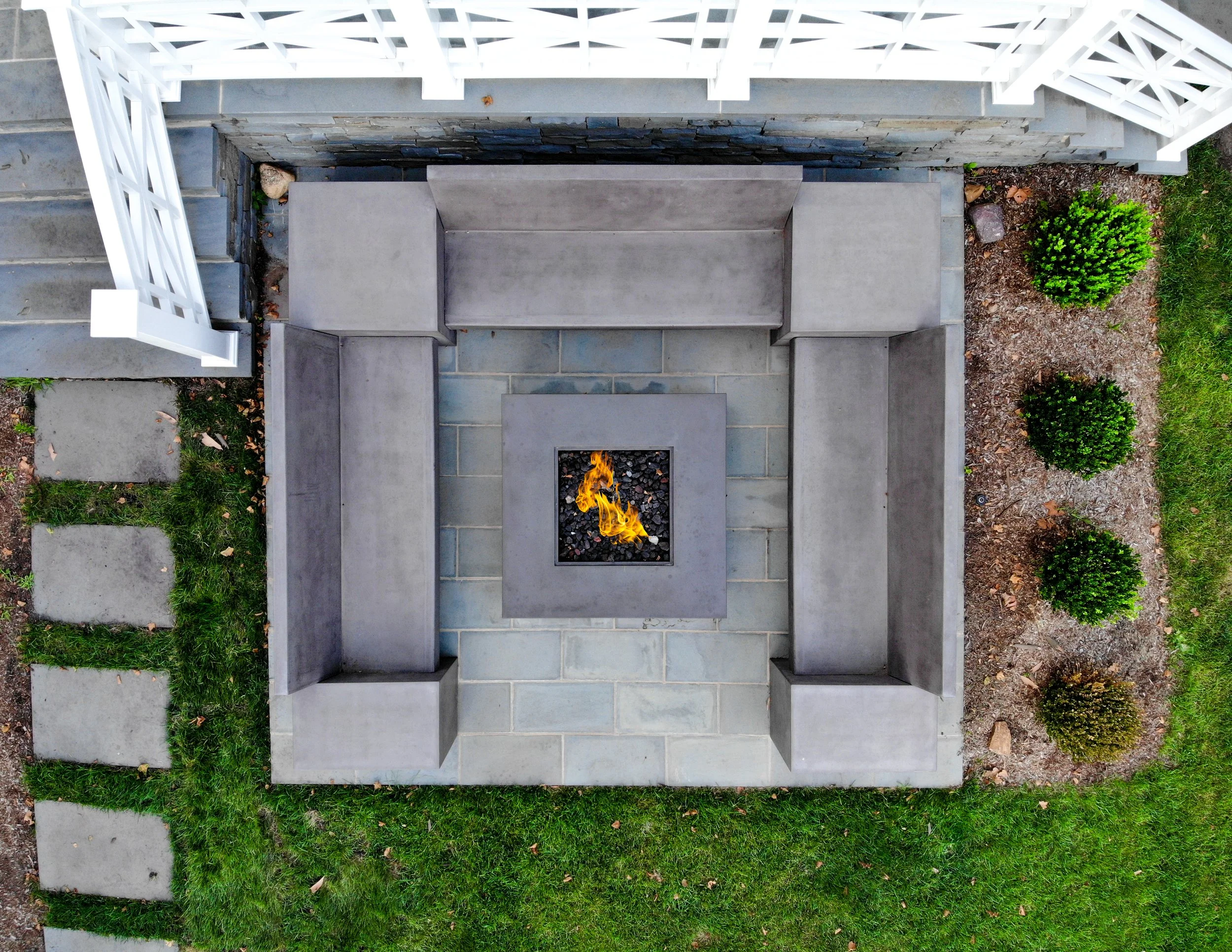 Top-down view of a patio with a fire feature at the center, surrounded by gray concrete bench seating, and bordered by a white fence, stepping stones on grass, and flower beds with three small green bushes.
