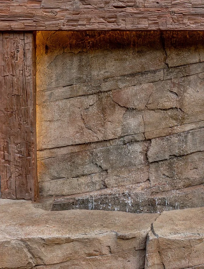 A close-up of a rock wall with a small water fountain at the bottom, framed by wooden beams and rocks.