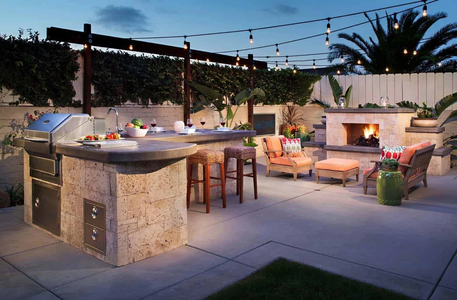 Outdoor patio with string lights, a built-in fireplace with a fire burning, a seating area with chairs and cushions, an outdoor kitchen with a countertop, tomatoes and wine glasses on the table, and lush greenery including palm trees and plants.