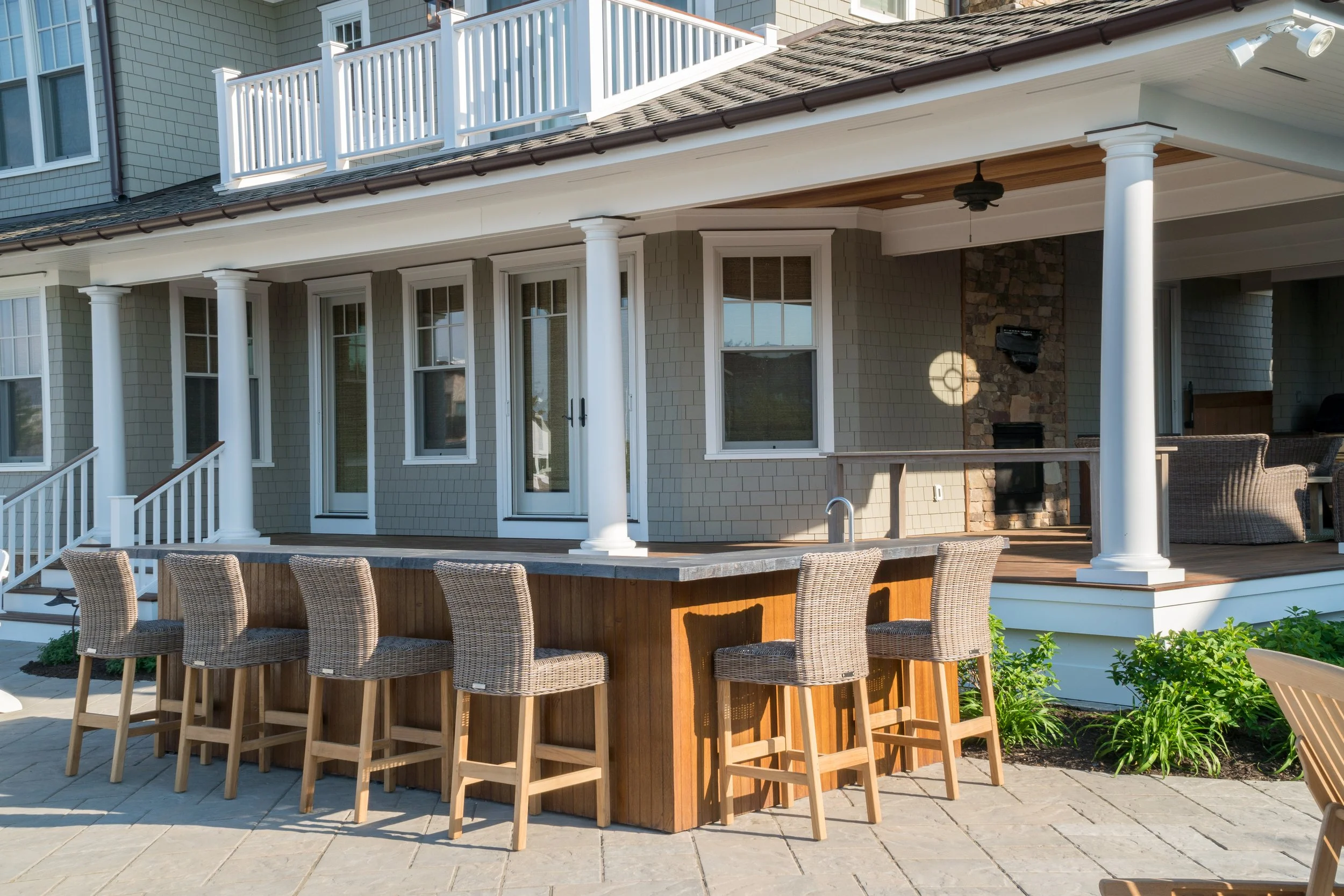 Backyard patio with an outdoor bar and six woven wicker chairs. The patio is attached to a house with light gray siding, white trim, and a covered porch with white columns. There is outdoor furniture and a grill in the background.