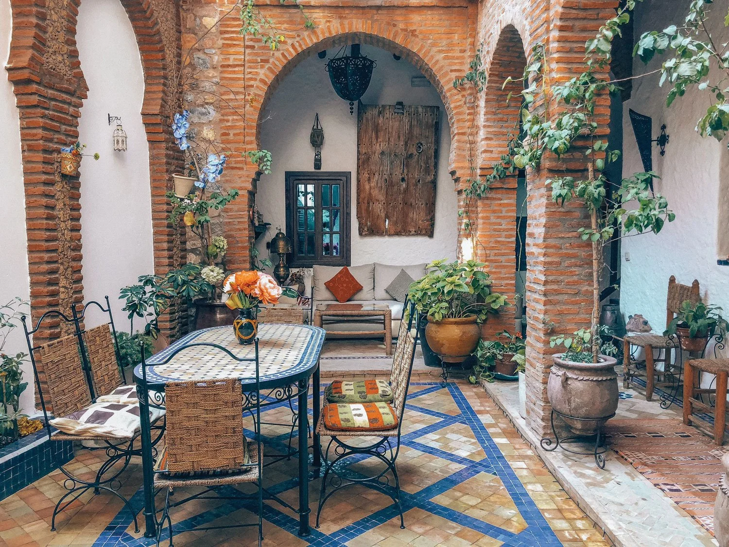 Indoor patio with brick arches, colorful tiled floor, wooden furniture, and potted plants.