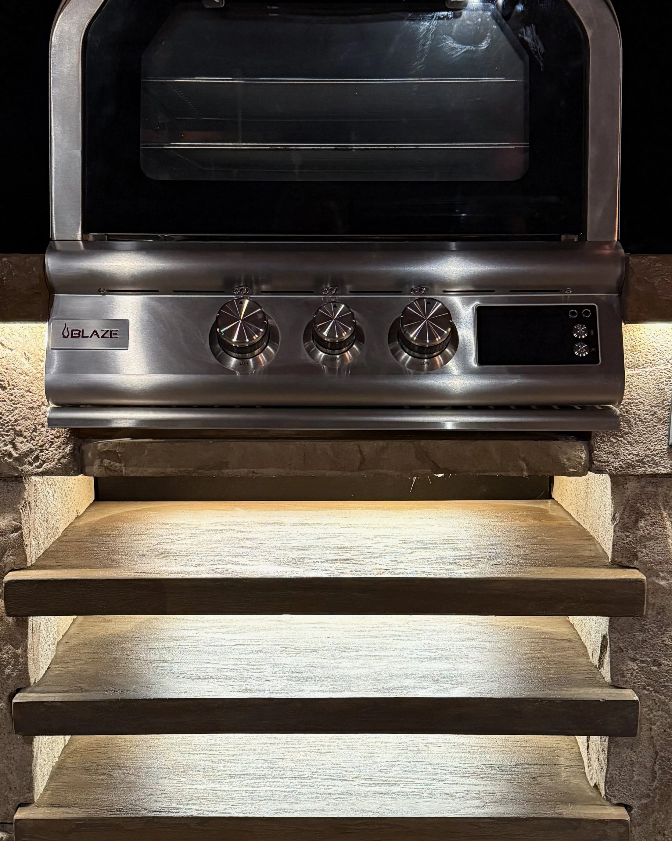 Stainless steel Blaze outdoor grill with three control knobs, built into a stone countertop, with illuminated wooden steps underneath.