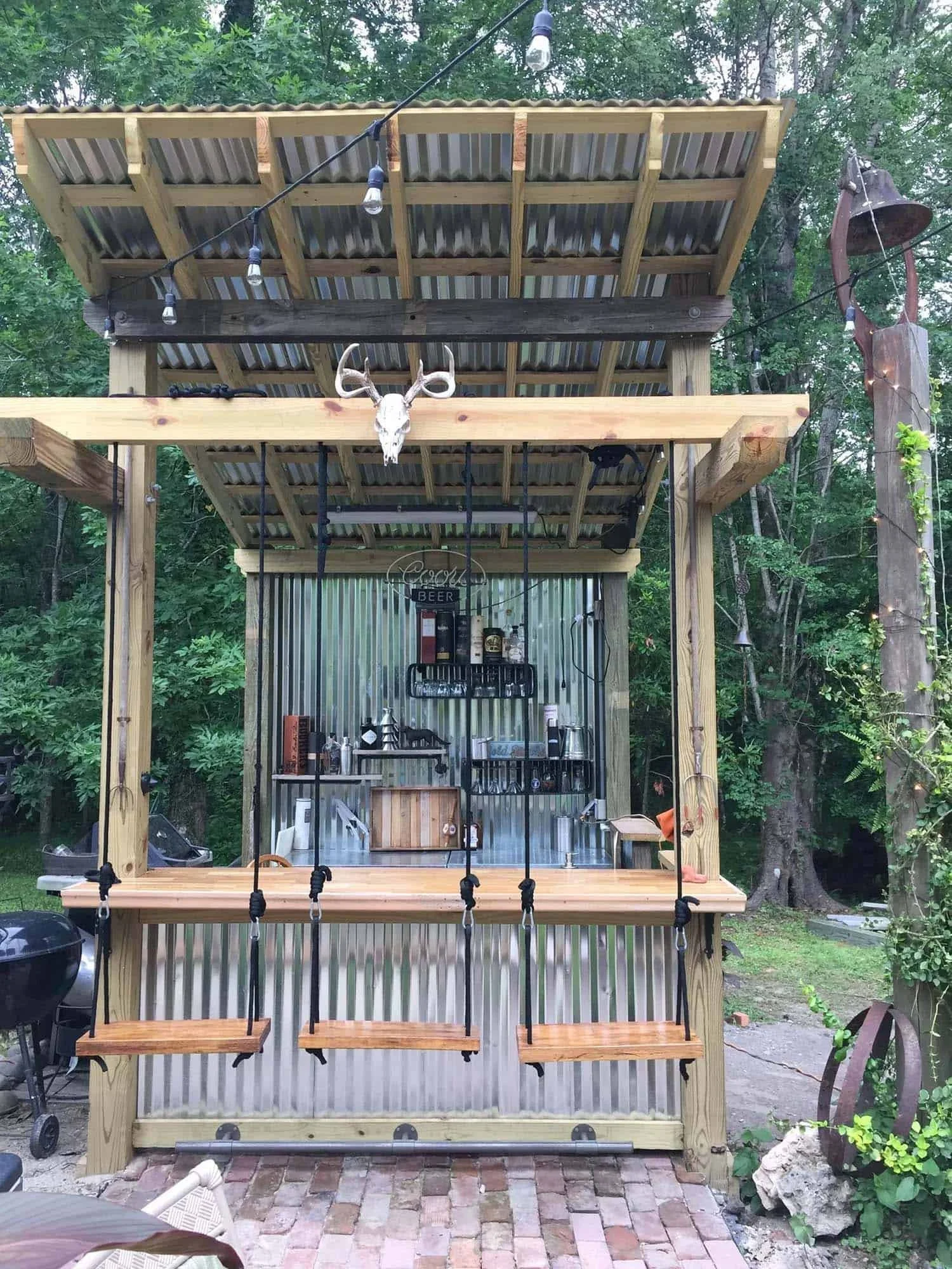 A backyard bar with a corrugated metal wall, hanging liquor bottles, a wooden countertop, and a swingset with three wooden swings in front.