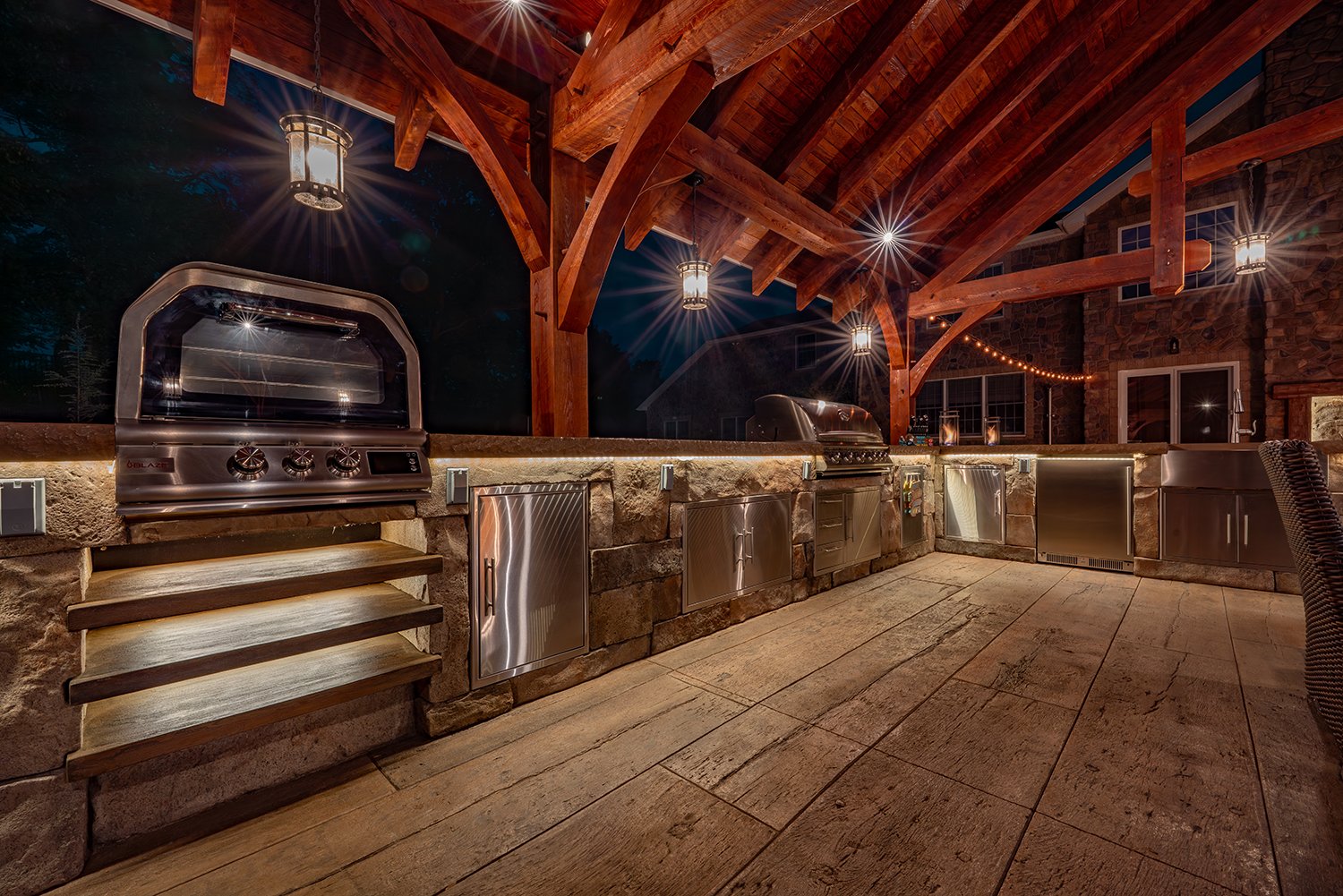 Luxury pavilion outdoor kitchen with stone counters, stainless steel grills, and wood-beam ceiling illuminated by pendant lights. The warm nighttime ambiance makes this backyard kitchen perfect for evening entertaining.
pavilion outdoor kitchen, nigh