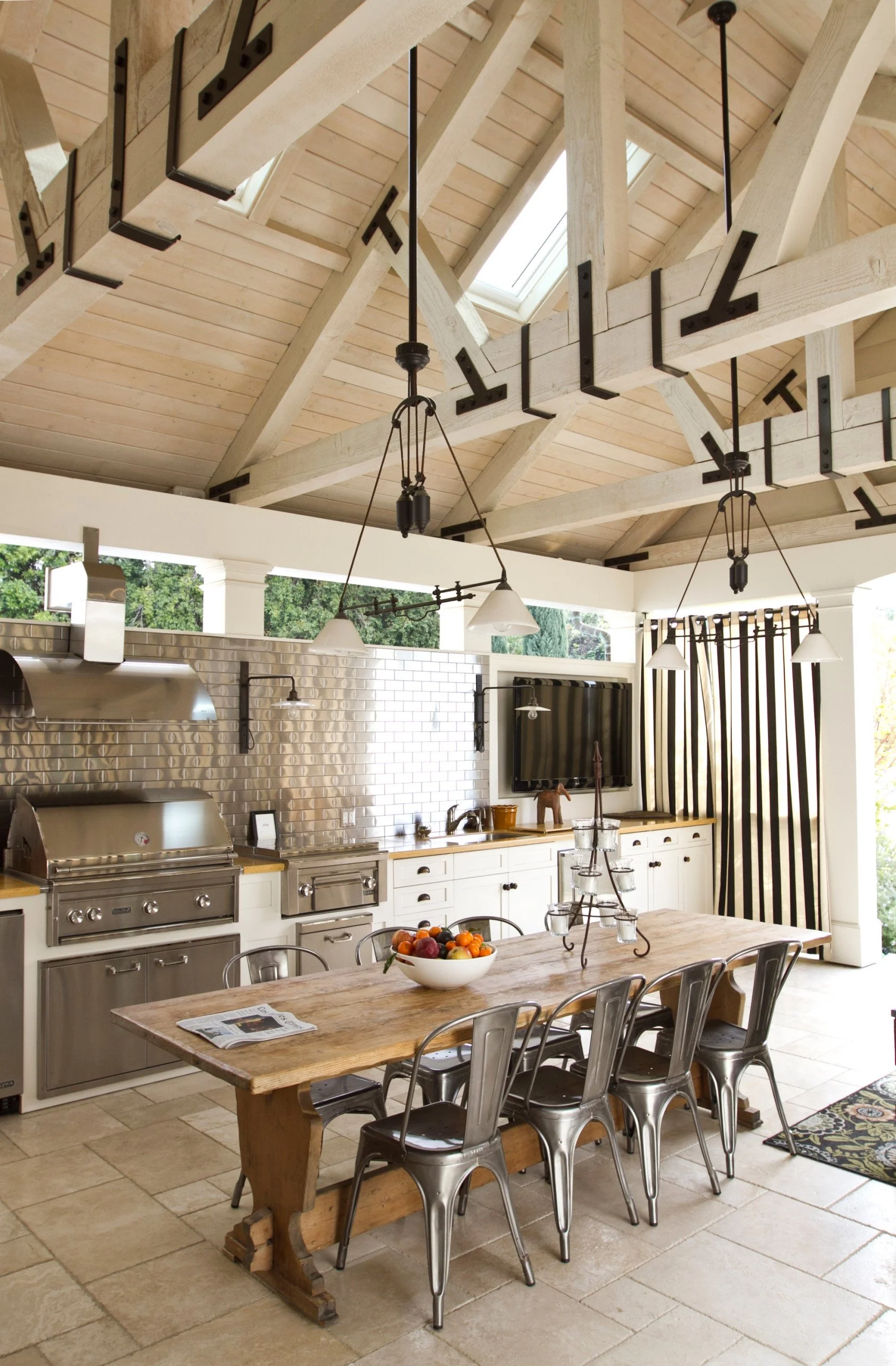 A rustic kitchen and dining area with a large wooden table, metal chairs, stainless steel appliances, white subway tile backsplash, a flat-screen TV, and a vaulted ceiling with skylights and hanging lights. A bowl of fruit is on the table.