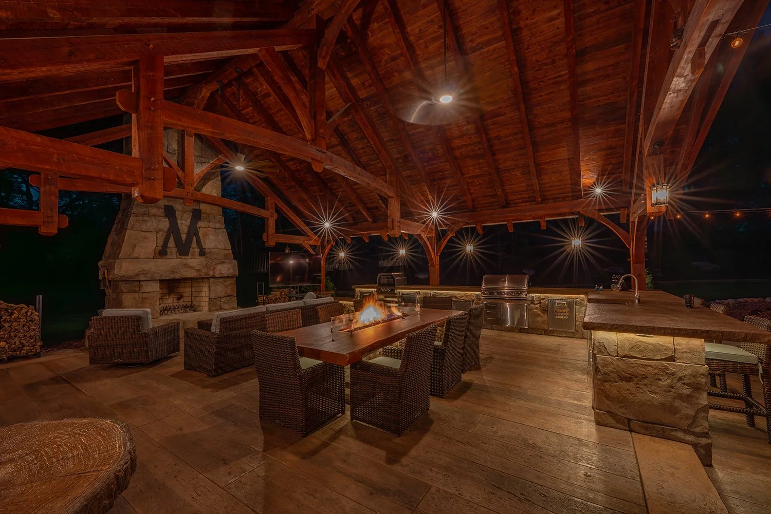 Built by Emil Gera. Rustic-style outdoor kitchen under a timber pavilion featuring a stone fireplace, dining fire table, wicker seating, and stainless steel grill. Perfect for cozy evenings and year-round backyard gatherings.
 rustic pavilion outdoor