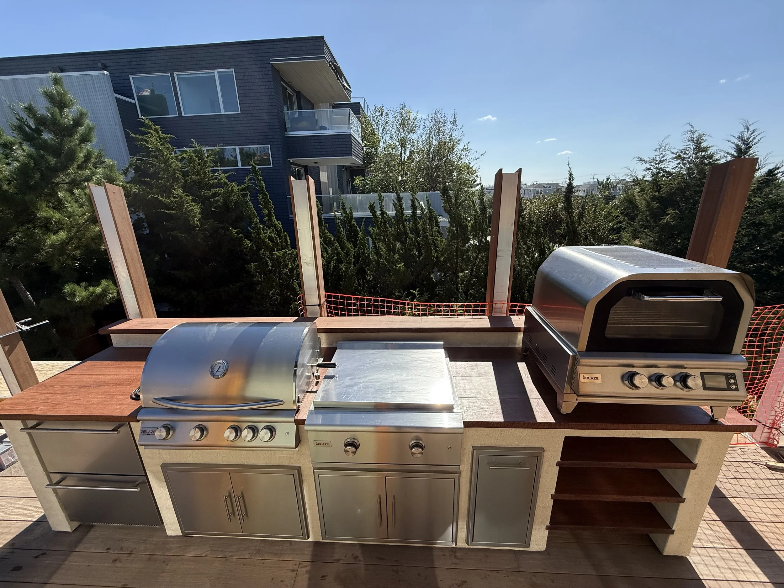 Outdoor kitchen with stainless steel grill, pizza oven, and countertop on a wooden deck, with trees and apartment building in the background.