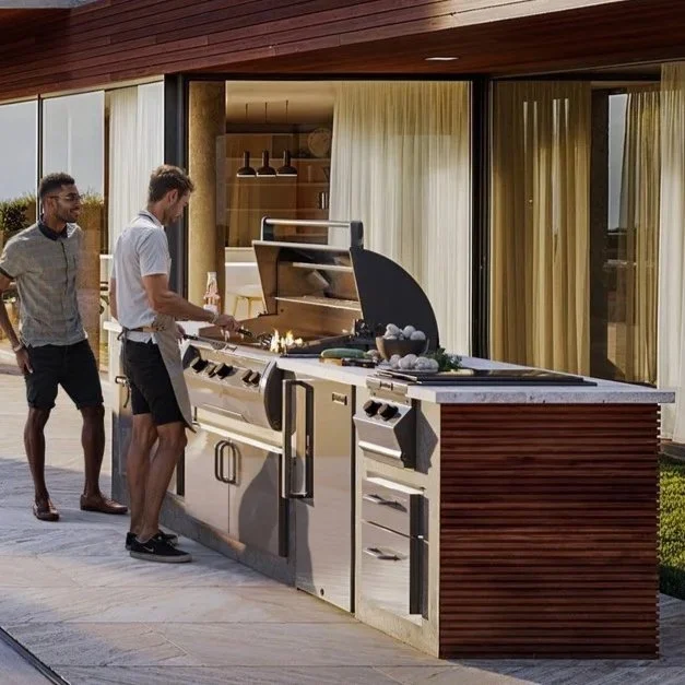 Two men are grilling food on a large outdoor barbecue with a modern house in the background.