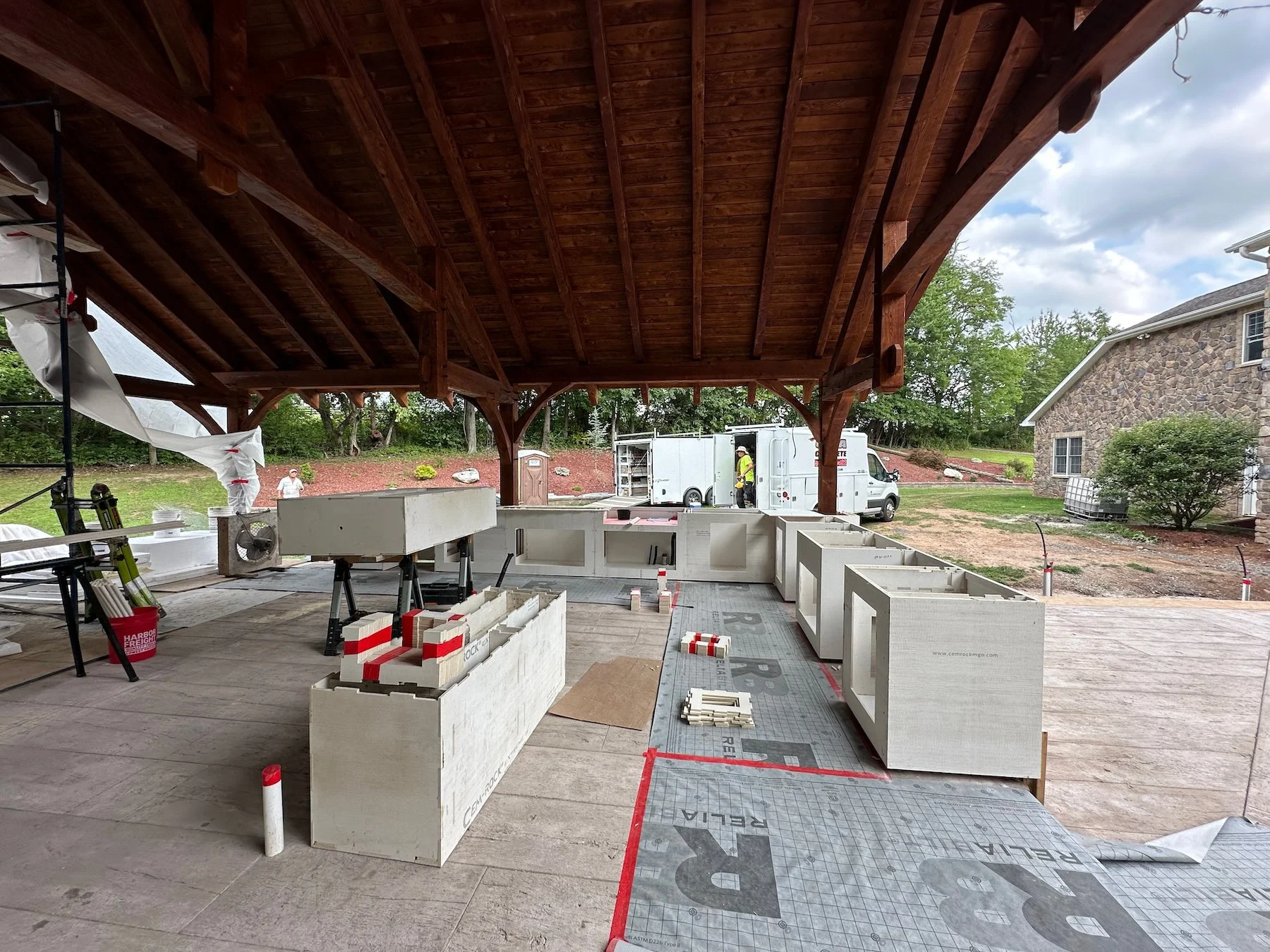Construction site of an outdoor kitchen under a wooden patio with building materials and a worker in a yellow safety vest near a white utility truck.