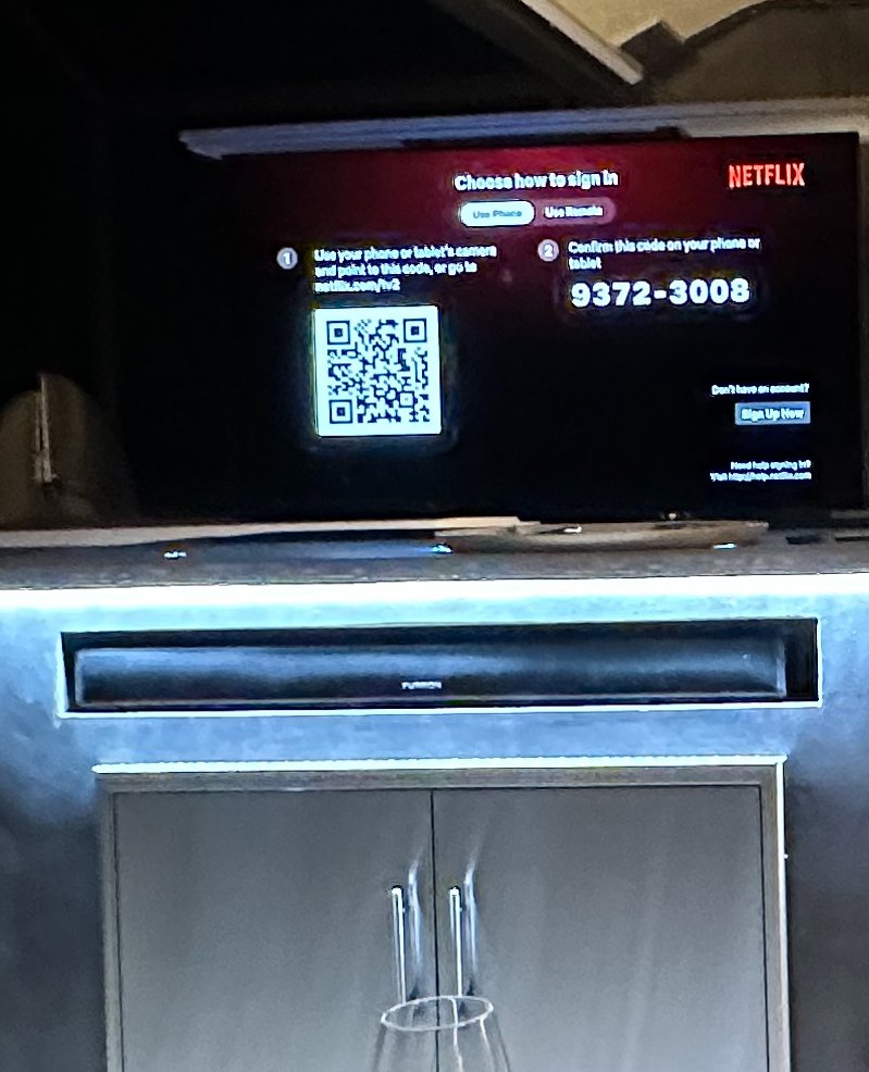 A television screen displays instructions for signing in to Netflix using a QR code and a phone number. The setup is on a cabinet with a short glass on top.