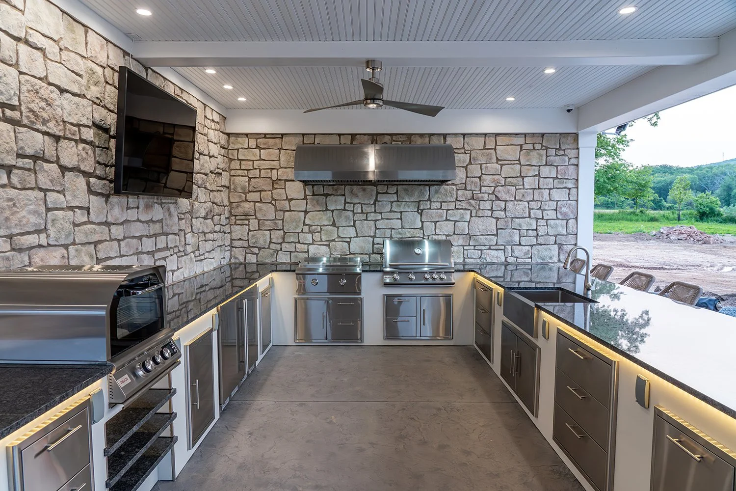 Built By Emil Gera. Modern outdoor kitchen with U-shaped stone veneer walls, stainless steel appliances, under-counter lighting, and granite countertops. Complete with ceiling fan, mounted TV, and dual built-in grills for the ultimate backyard cookin