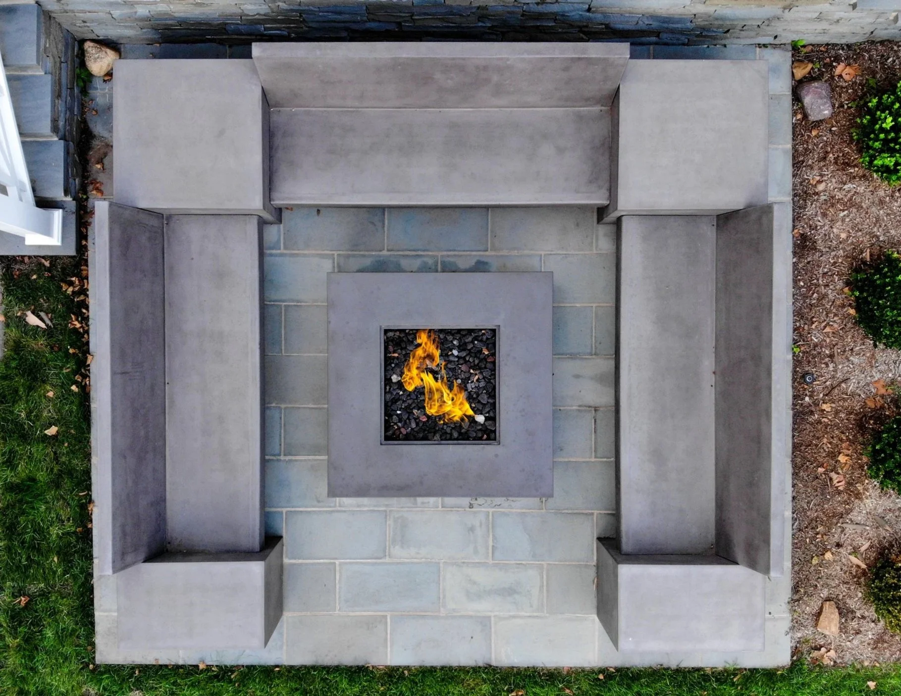 Overhead view of a concrete outdoor fire pit with a small flame, surrounded by built-in concrete seating and a stone patio, with grass, plants, and dirt edging in the background.