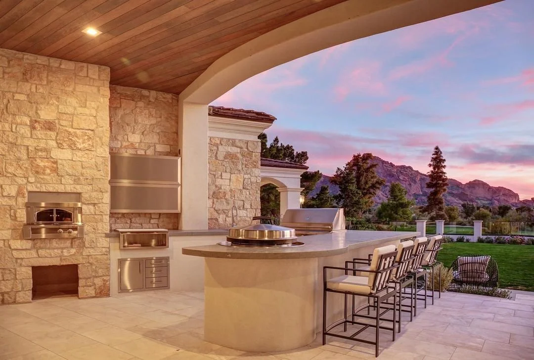 Outdoor kitchen with stone walls, stainless steel appliances, bar counter with four chairs, mountains and trees in the background, pink and purple sky at sunset.