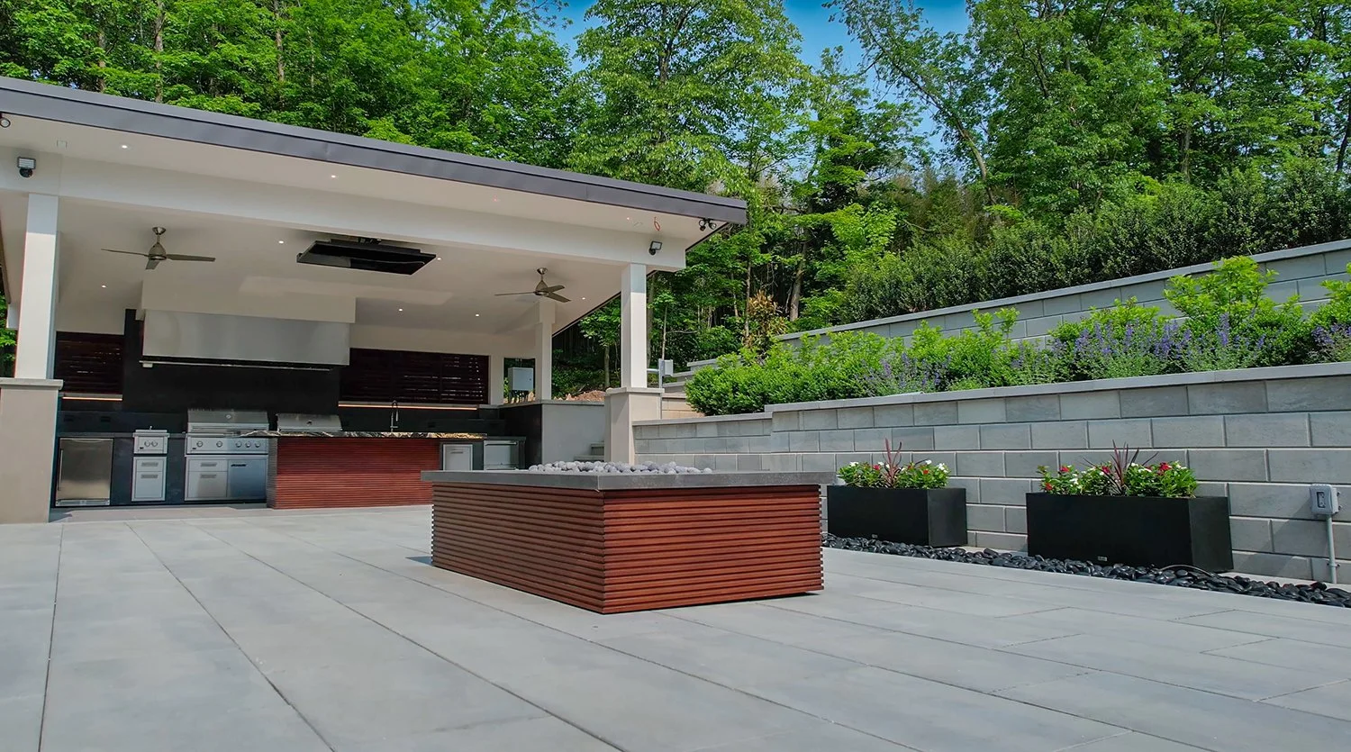 Contemporary covered outdoor kitchen with sleek appliances, modern pergola roof, and a central fire pit table. Surrounded by retaining walls and landscaping, this backyard design blends modern style with outdoor entertaining.
modern outdoor kitchen, 