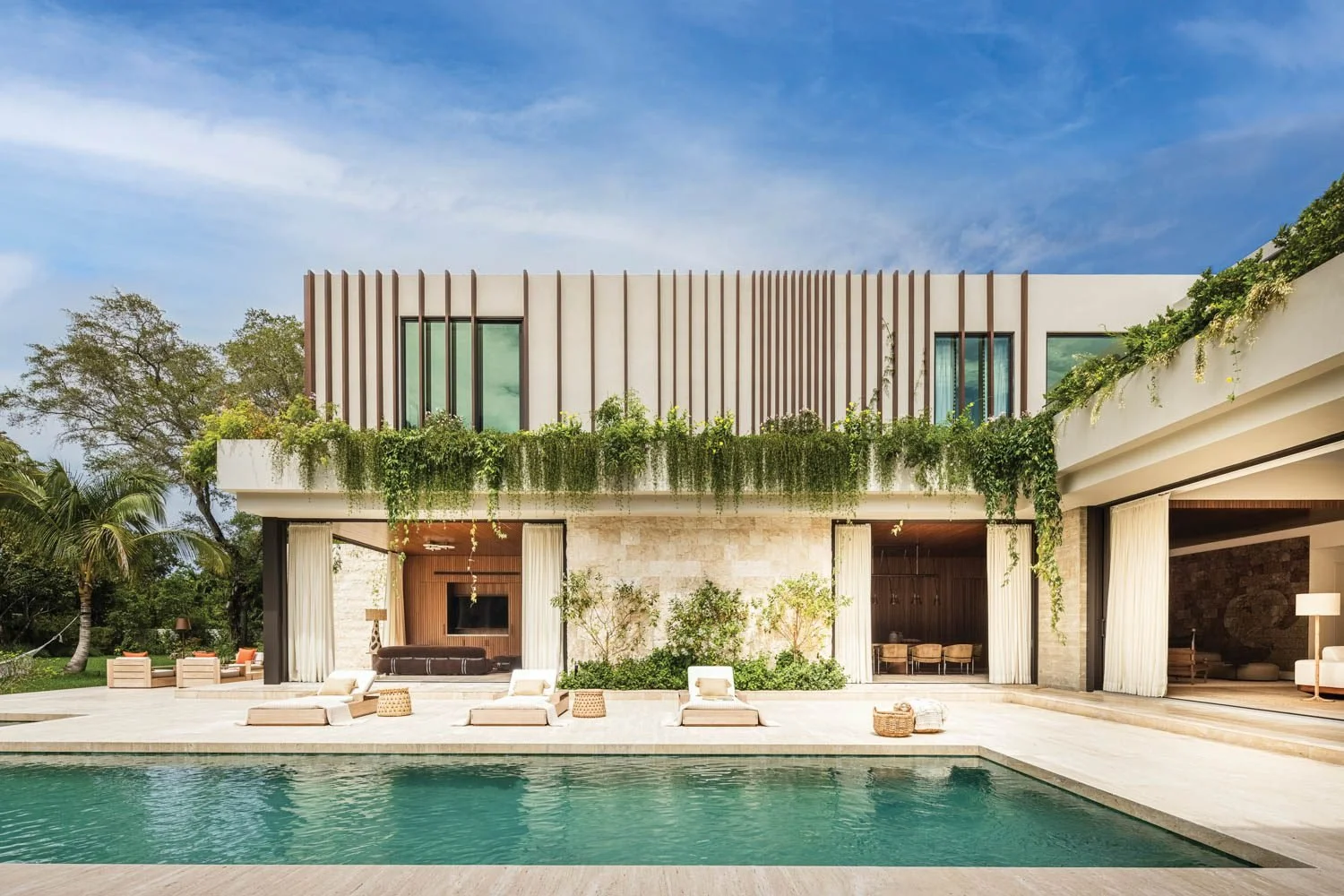Modern house with a swimming pool, outdoor lounge area, and lush greenery under a blue sky.