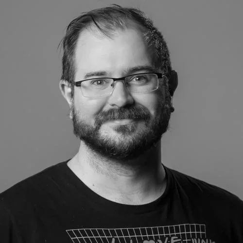 Black and white portrait of a man with glasses, a beard, and short hair, wearing a T-shirt against a plain background.