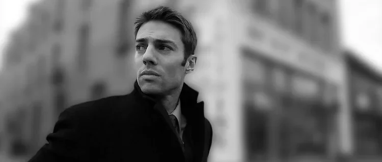 A young man with short hair dressed in a dark coat looking to the side in an urban setting, black and white image.